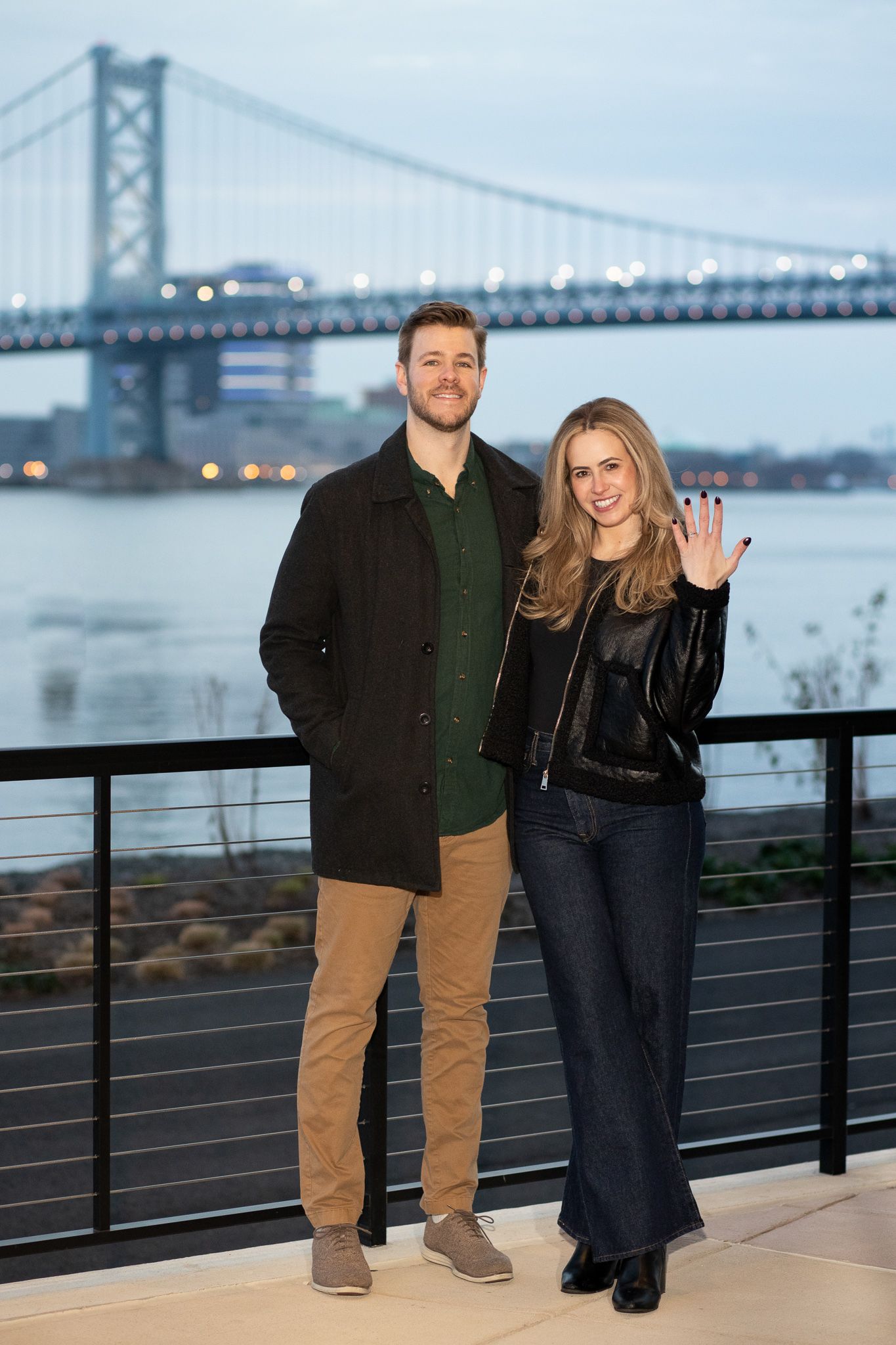 A couple poses together on a waterfront terrace with the Ben Franklin Bridge visible in the background at dusk.