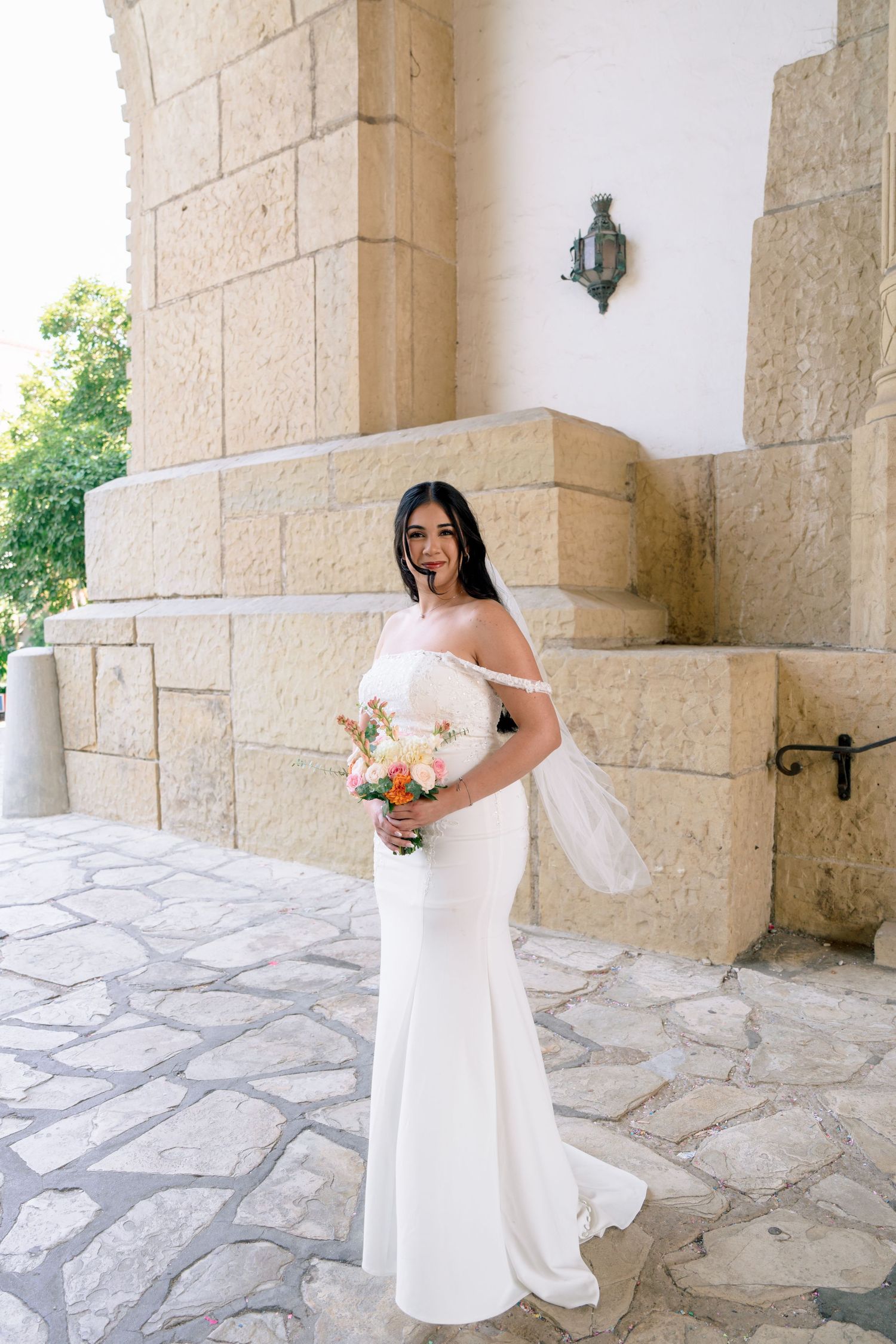 A bride in a white off-shoulder wedding dress poses elegantly in a stone archway courtyard.