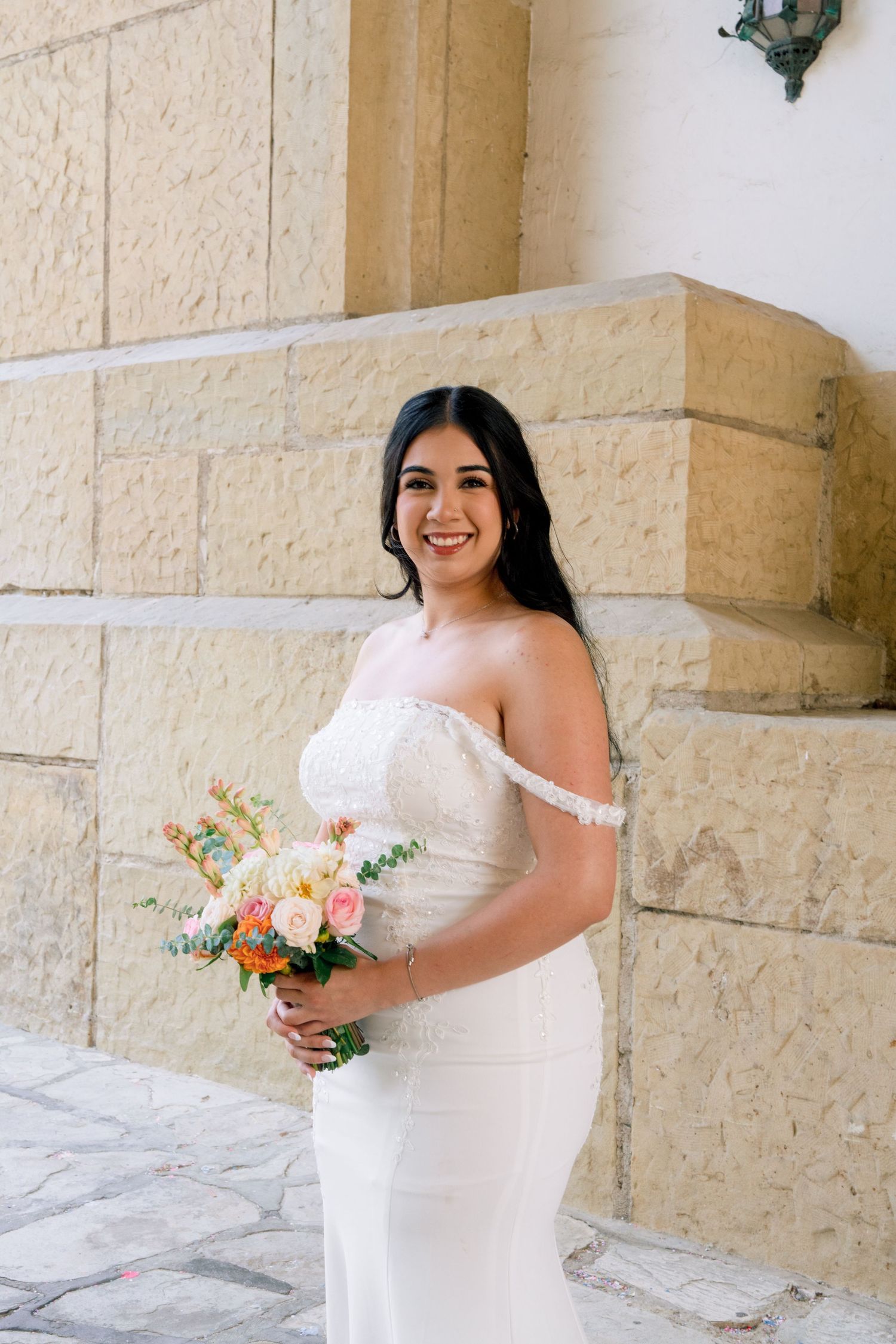 A bride in an off-shoulder wedding dress holds a colorful bouquet against a stone wall.