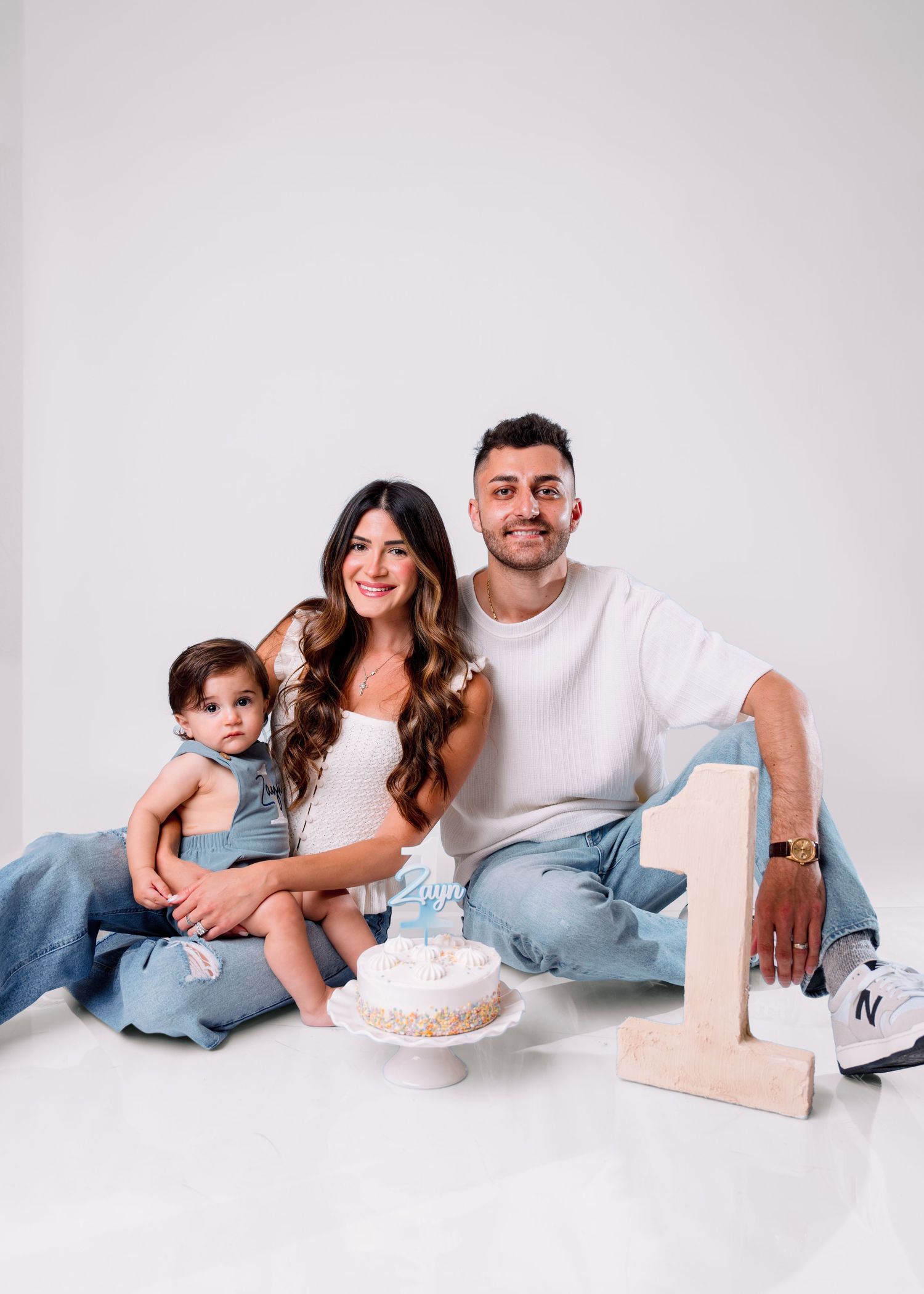 Family photo with parents and baby celebrating first birthday with cake and wooden number one prop.