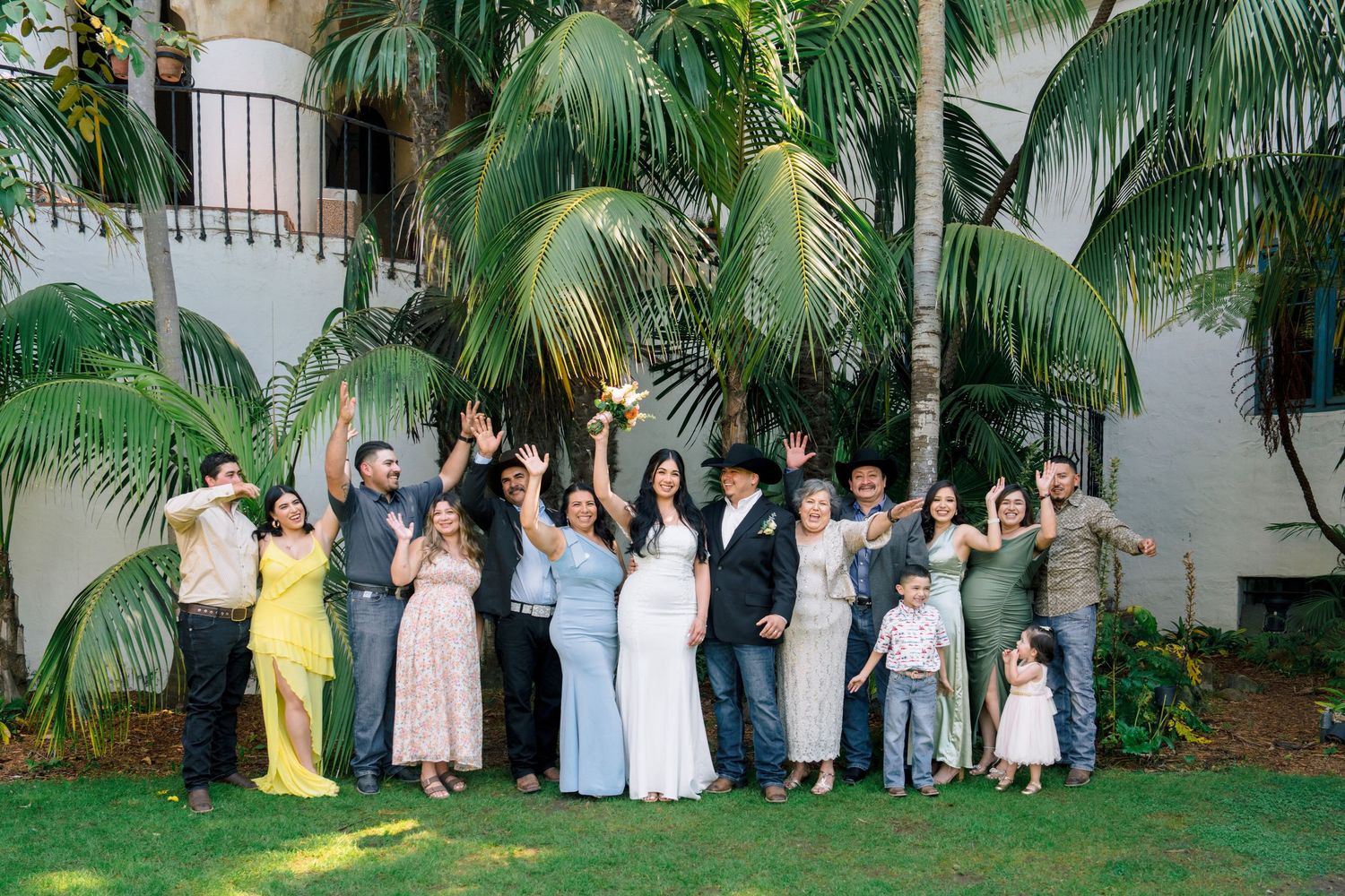 Wedding party members gather for photos under palm trees on a sunny day.