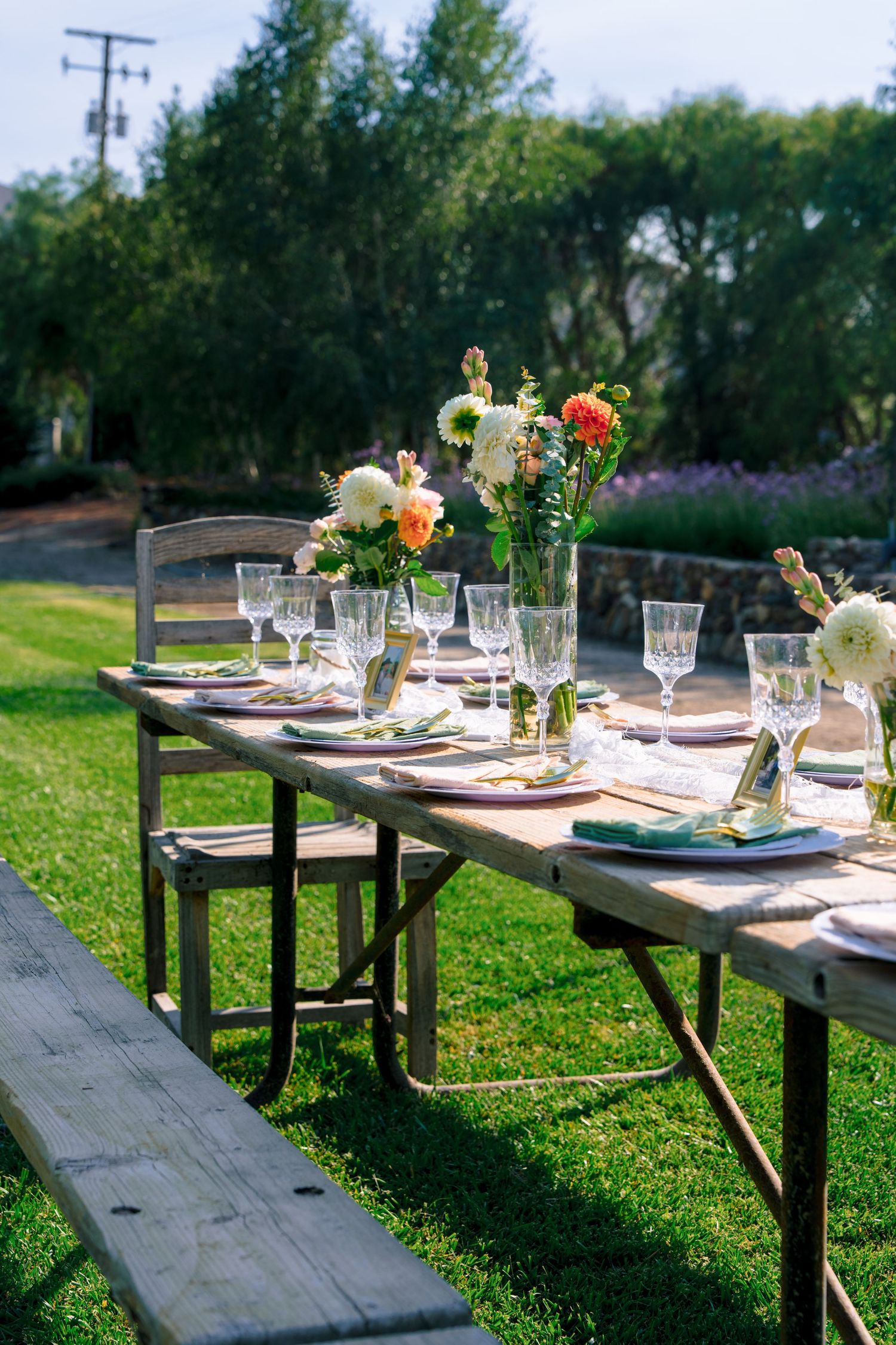 Elegant outdoor table setting with floral centerpieces and wooden benches on a sunny lawn.
