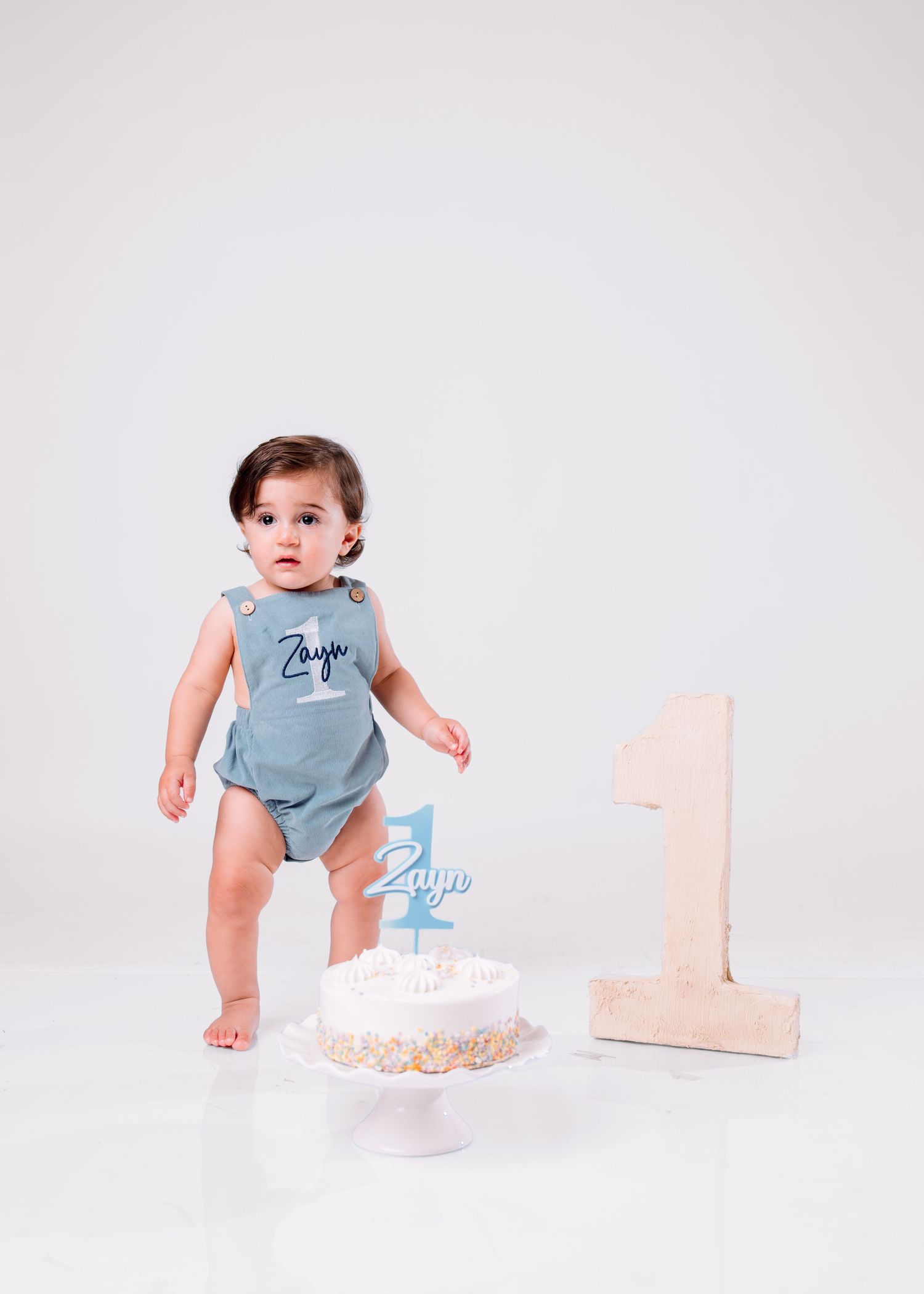Baby boy named Zayo stands by his first birthday cake and number one decoration, wearing a blue romper.
