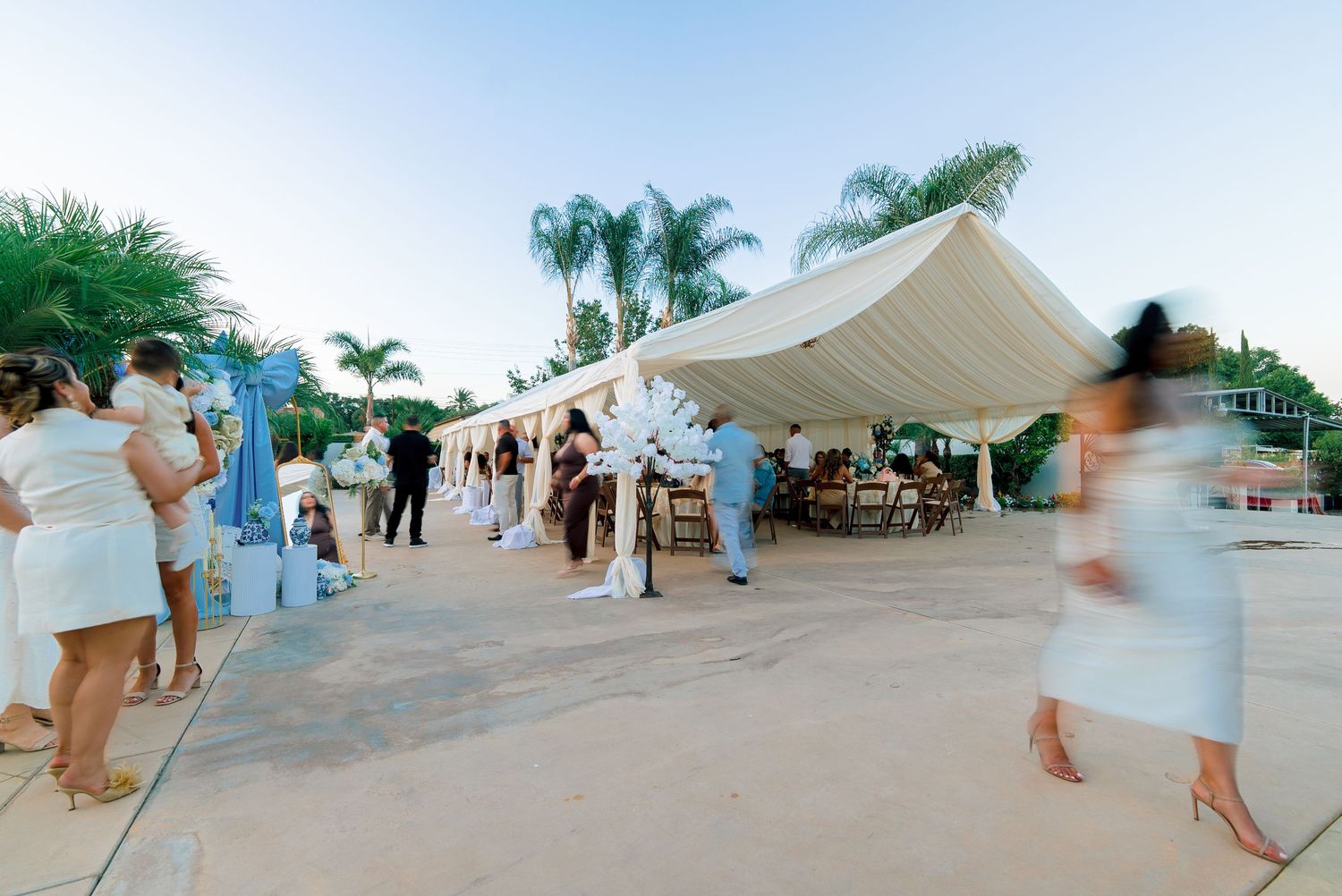 Outdoor wedding reception under a white tent with guests dressed in white, palm trees, and elegant floral decorations at dusk.