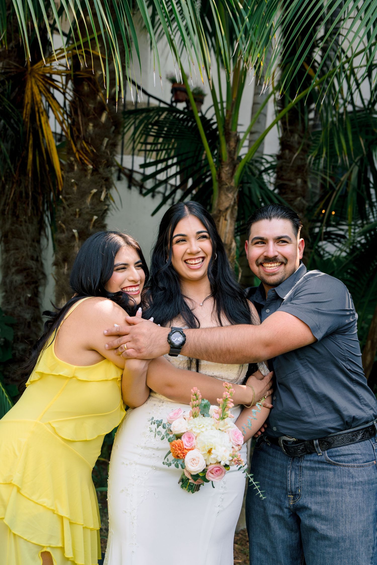 Wedding party members share laughs and hugs in front of palm trees.