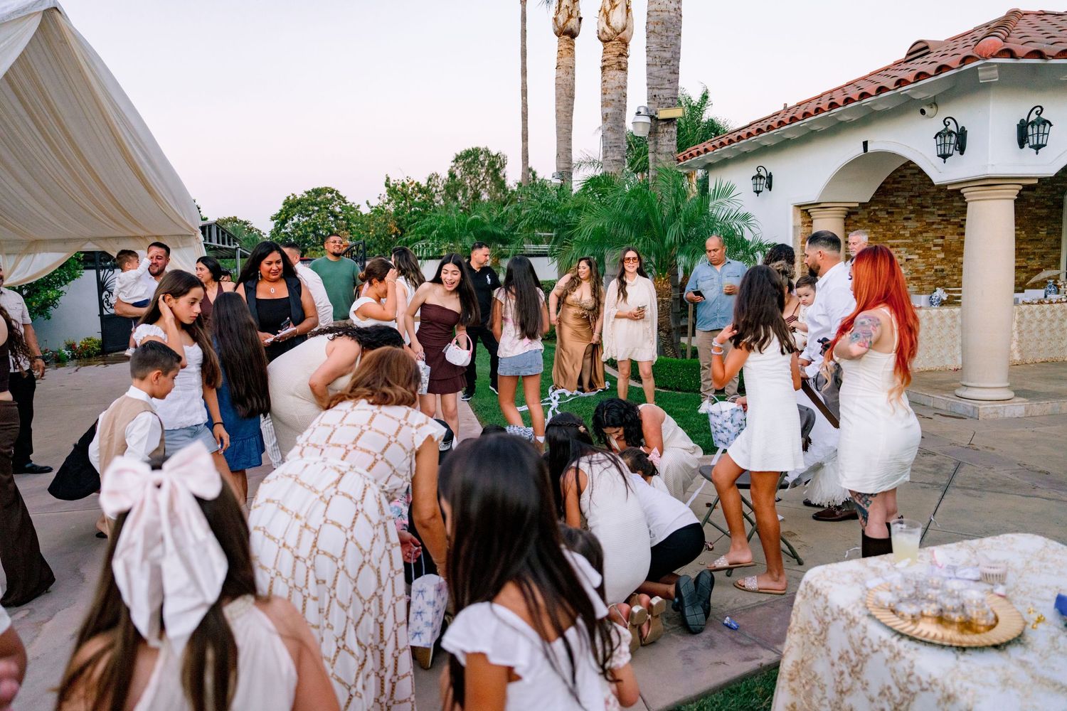 Guests in white attire gather outdoors at an elegant garden party near a Spanish-style villa at dusk.