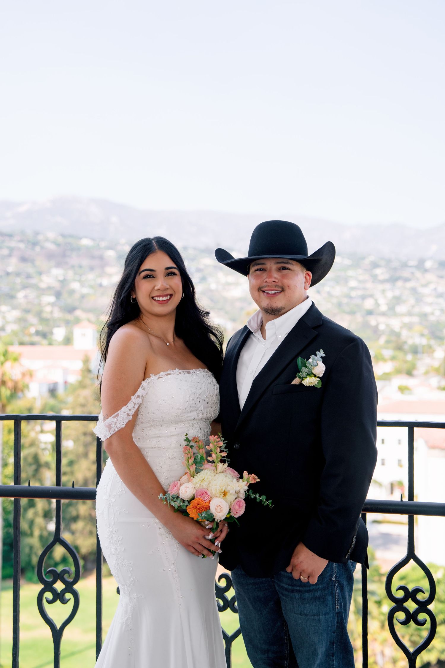 Wedding couple poses on a balcony overlooking a scenic city vista.