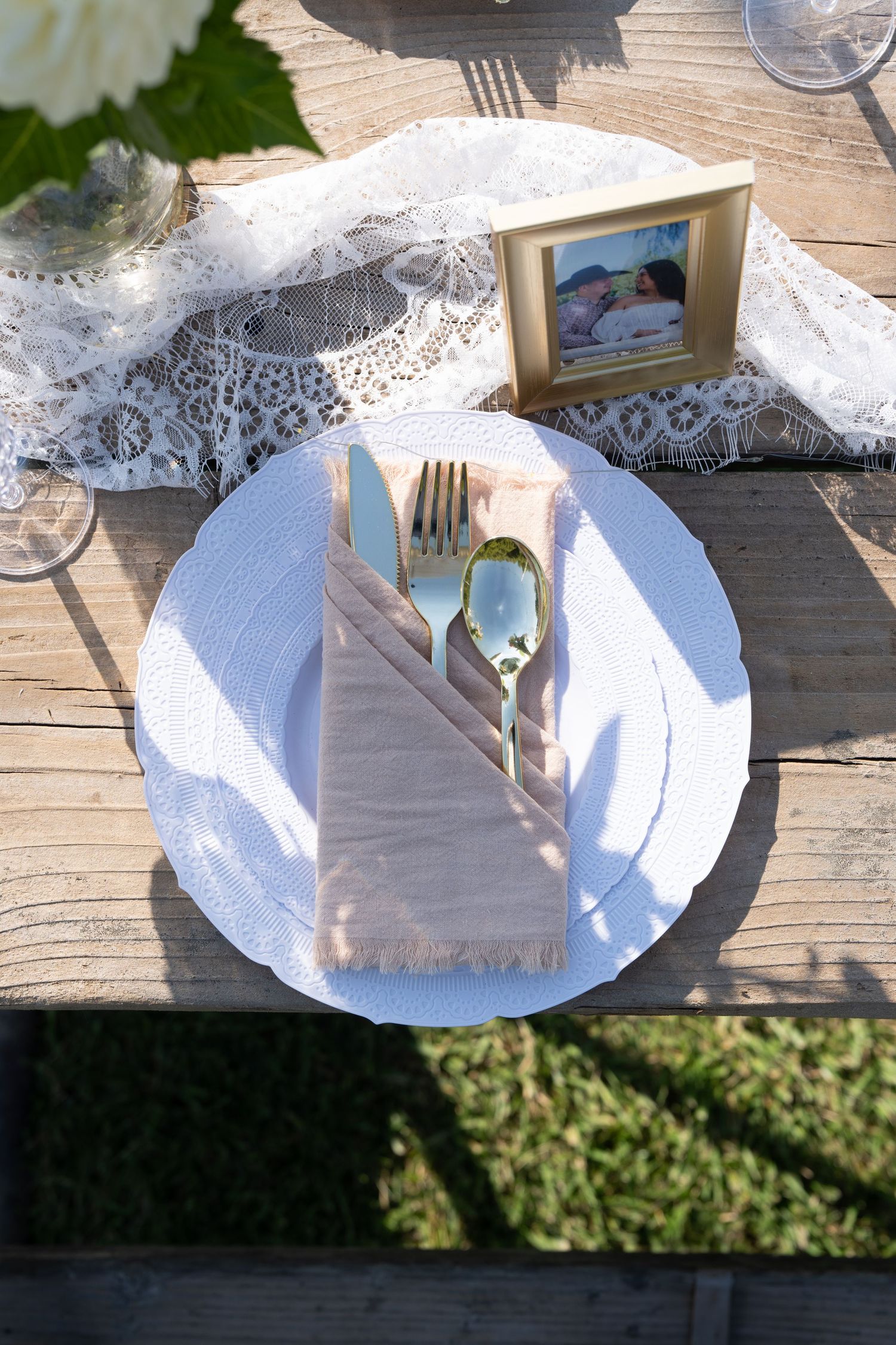 Elegant white place settings with lace details and gold frames on wooden tables outdoors.