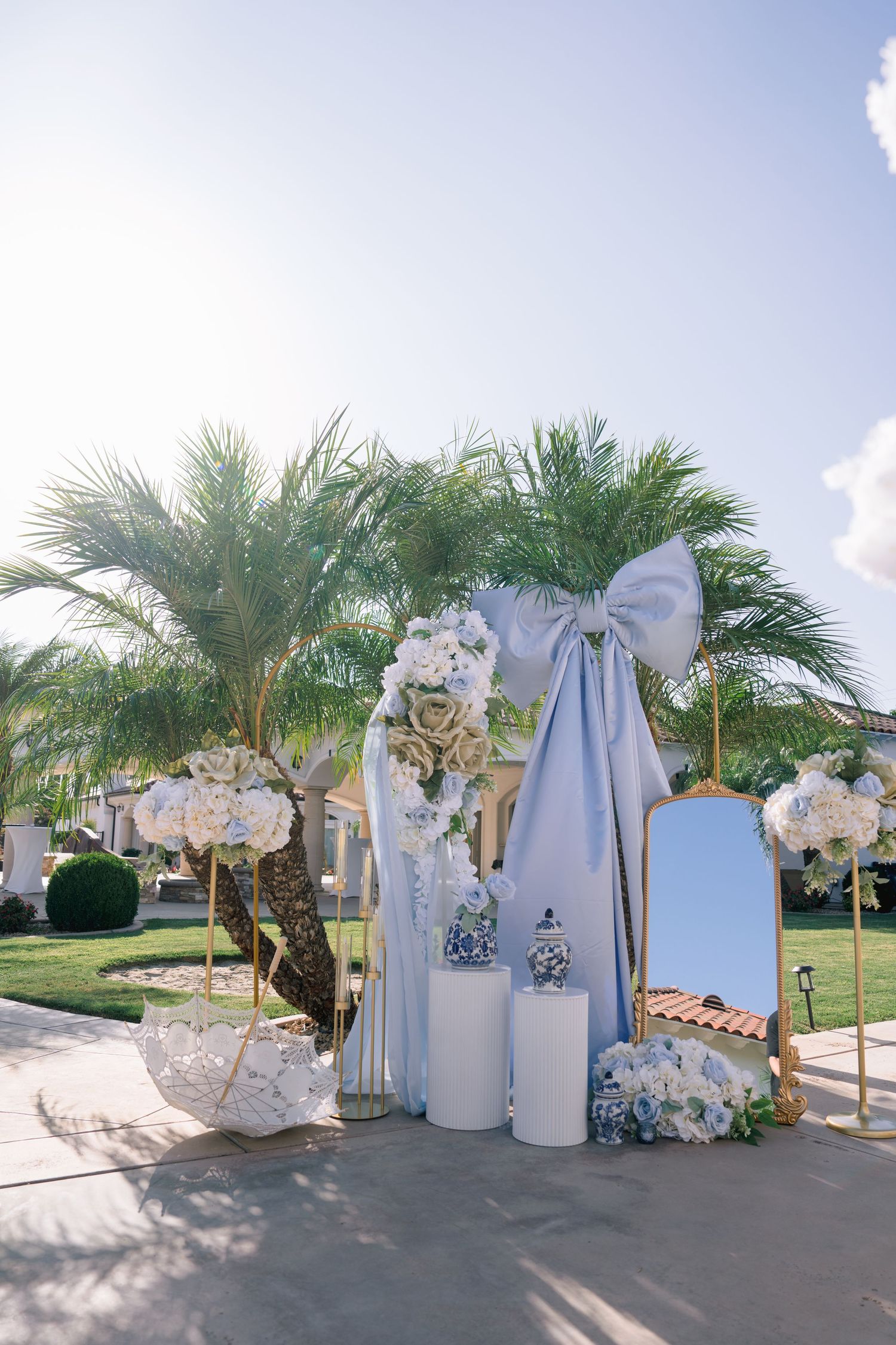 Elegant outdoor wedding arch with light blue bow, white flowers, mirror, and palm trees under a bright sky.