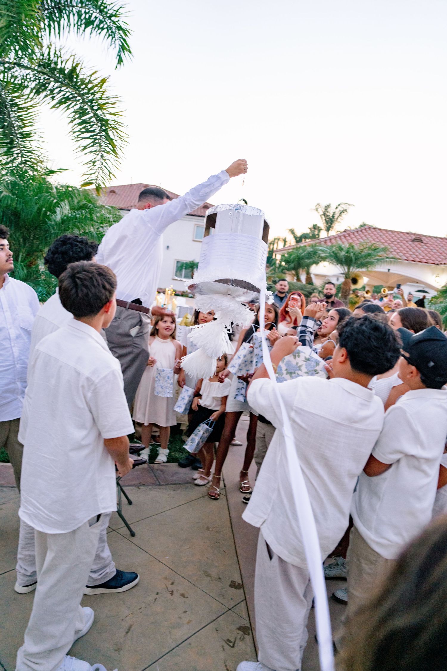 Group of people in white outfits breaking a pinata at an outdoor party near a Spanish-style home.