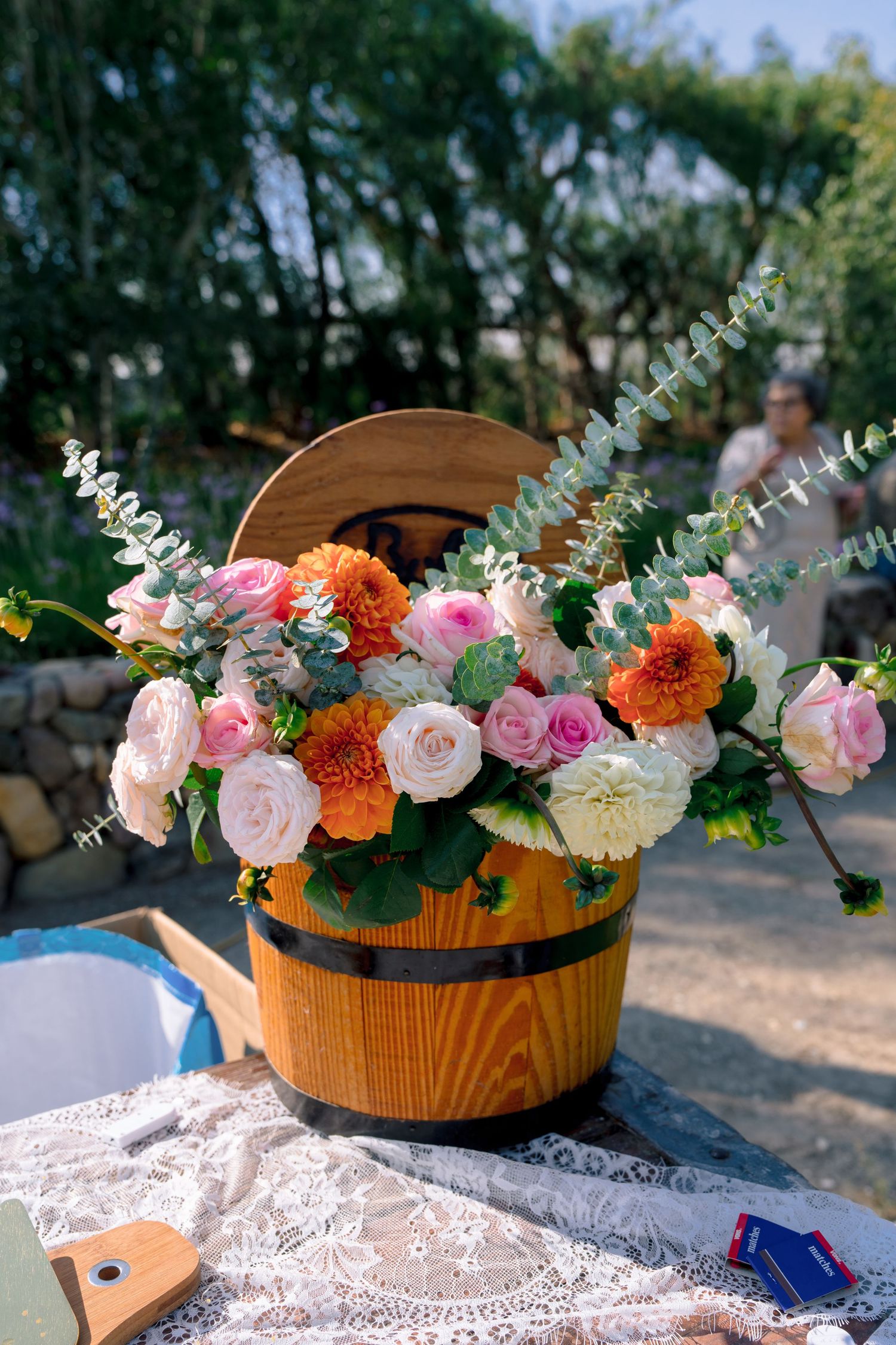 Colorful floral arrangement with pink roses and orange flowers in a rustic wooden bucket on a lace tablecloth.