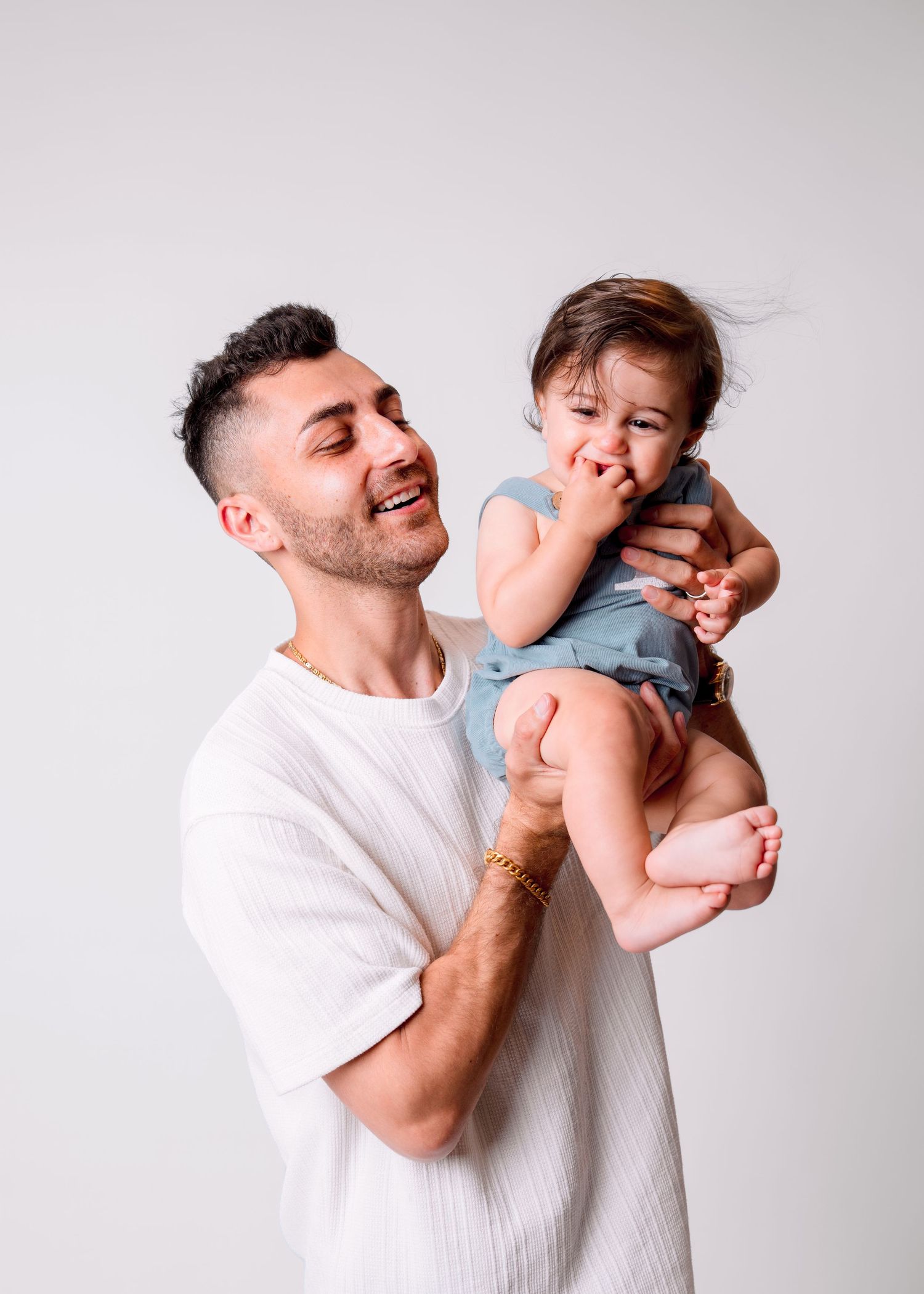 A smiling man holds a happy baby in a blue romper against a white background.