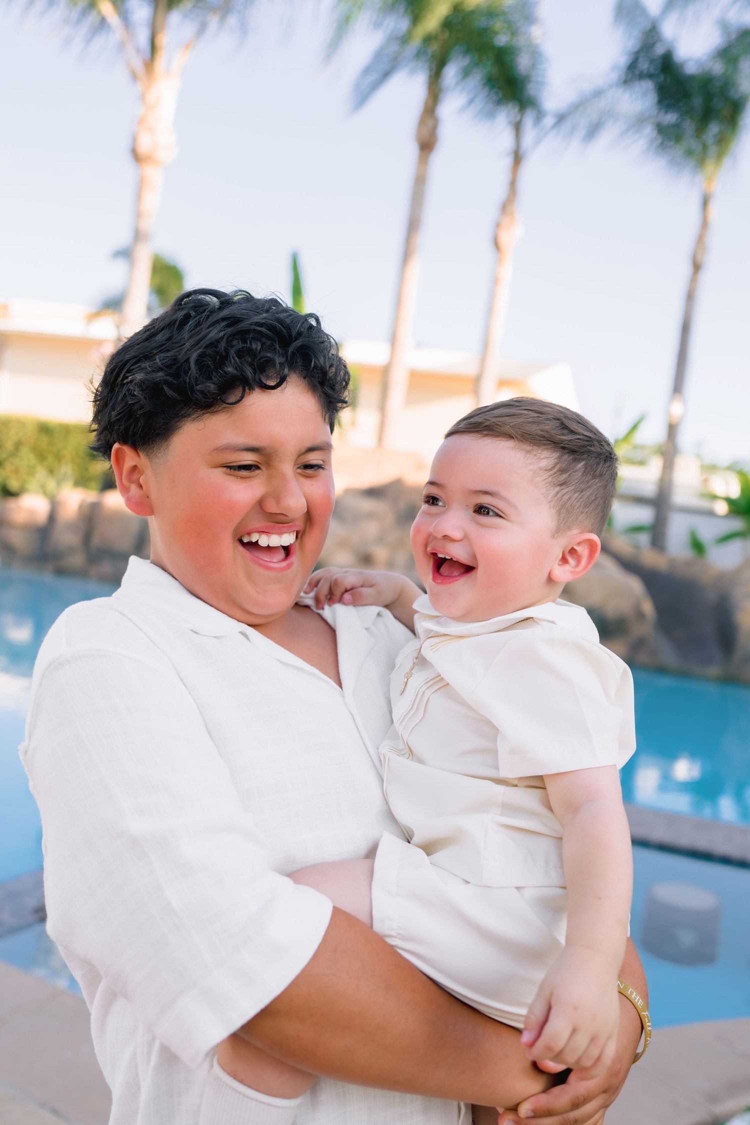 A laughing woman in white holds a joyful baby by a pool with palm trees in the background.