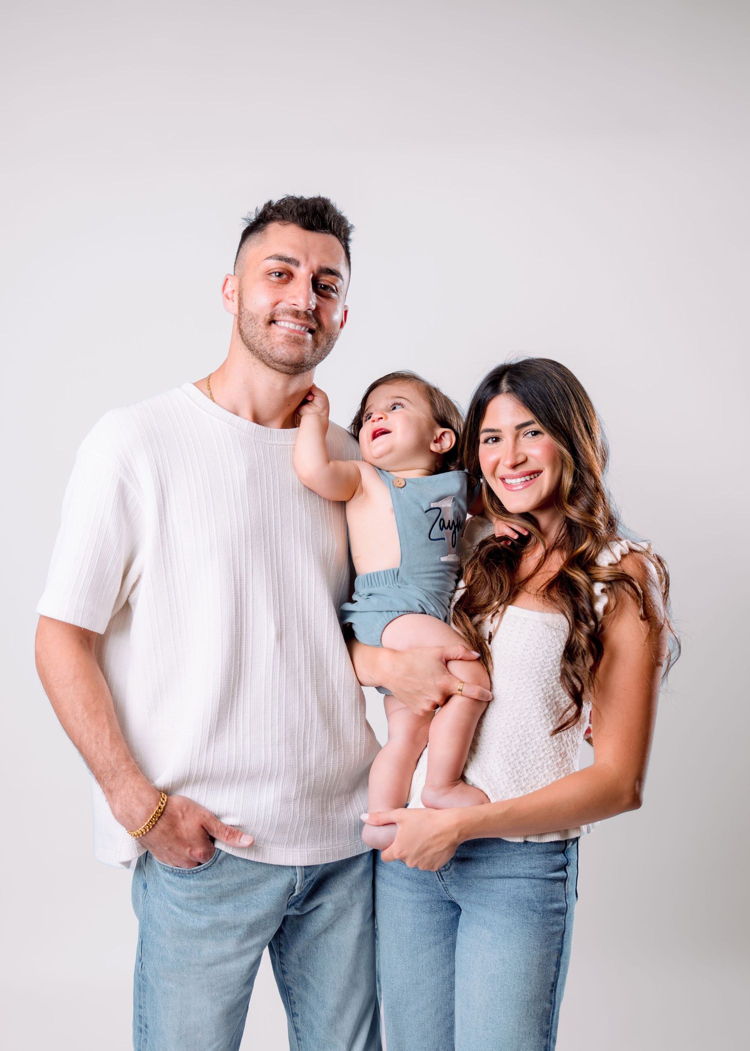A young family poses together, two parents holding a smiling baby dressed in blue, against a white studio background.