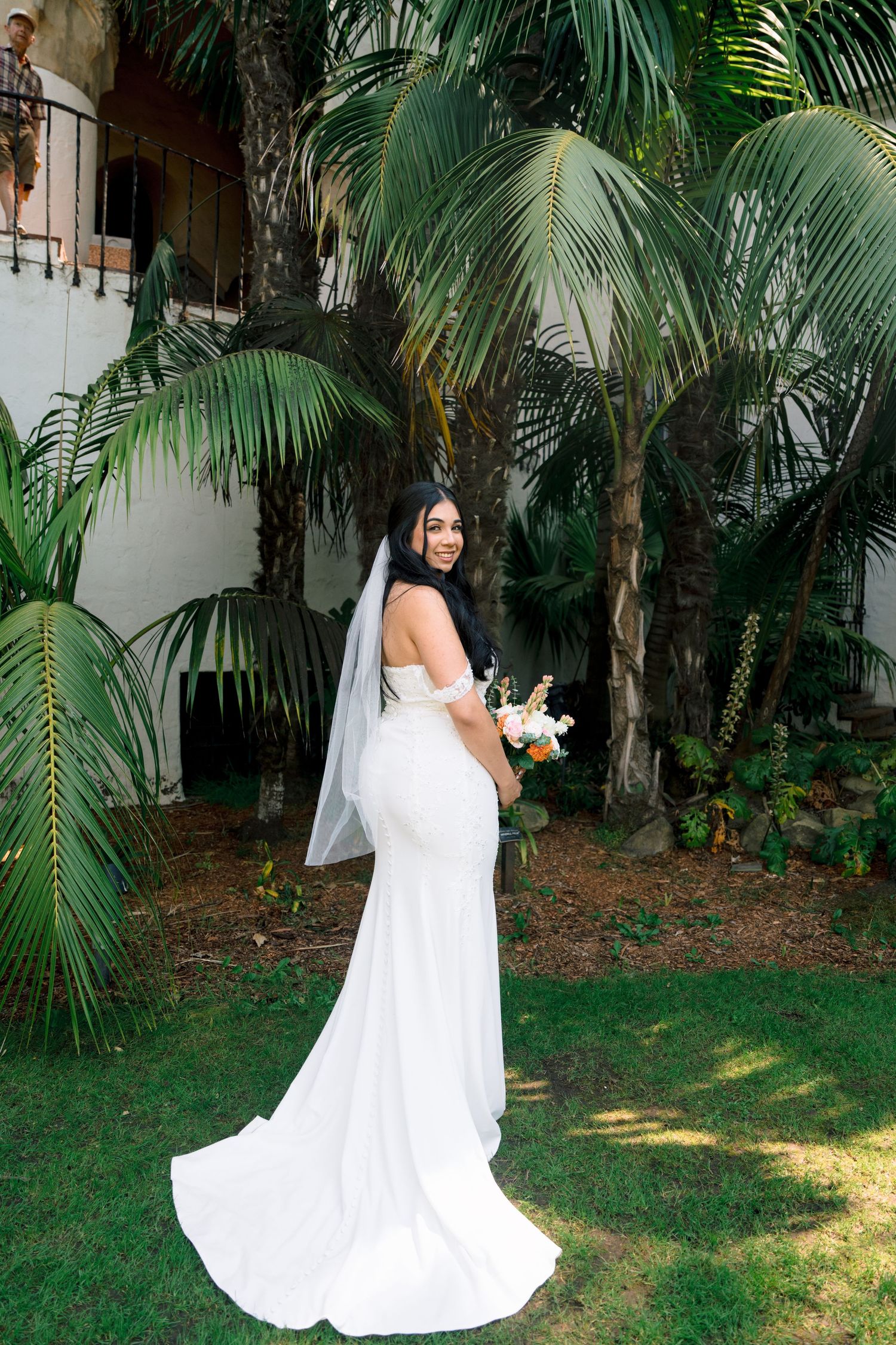 Bride in flowing white dress poses in a tropical garden setting with palm trees.