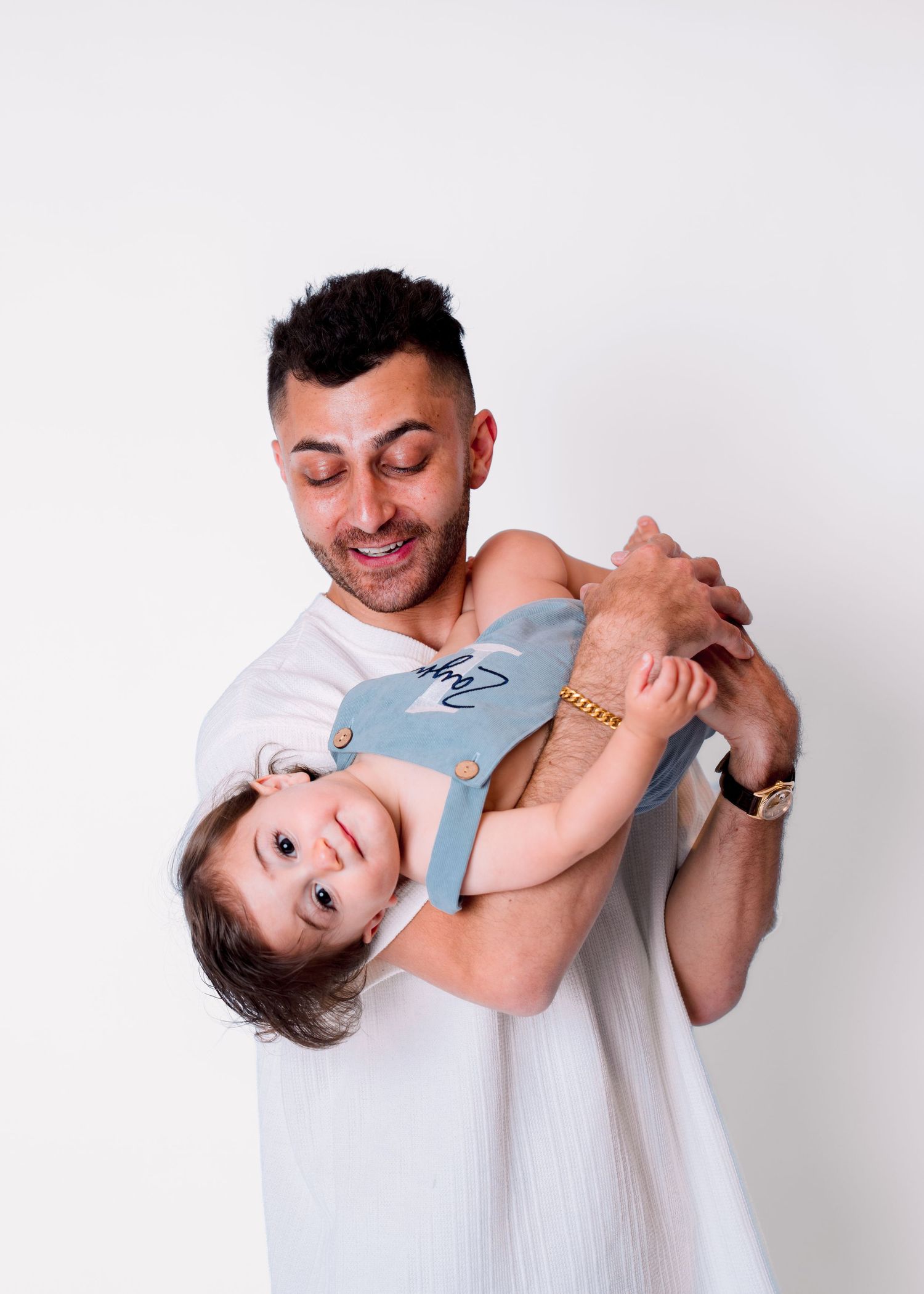 A smiling man in a white shirt joyfully holds a toddler in blue overalls against a white background.