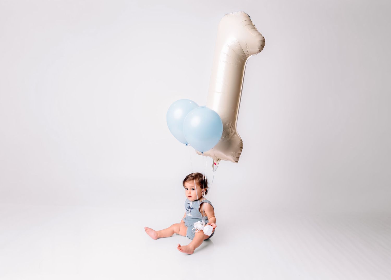 Baby boy in blue romper sits on white floor holding a light blue balloon and number 1 foil balloon for first birthday.