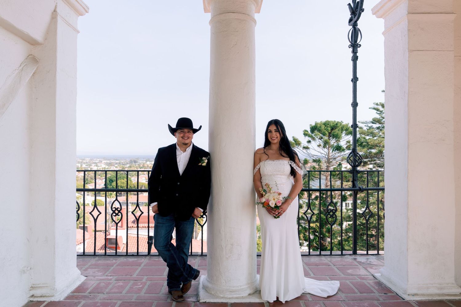 Wedding couple standing between white columns in an outdoor colonnade.