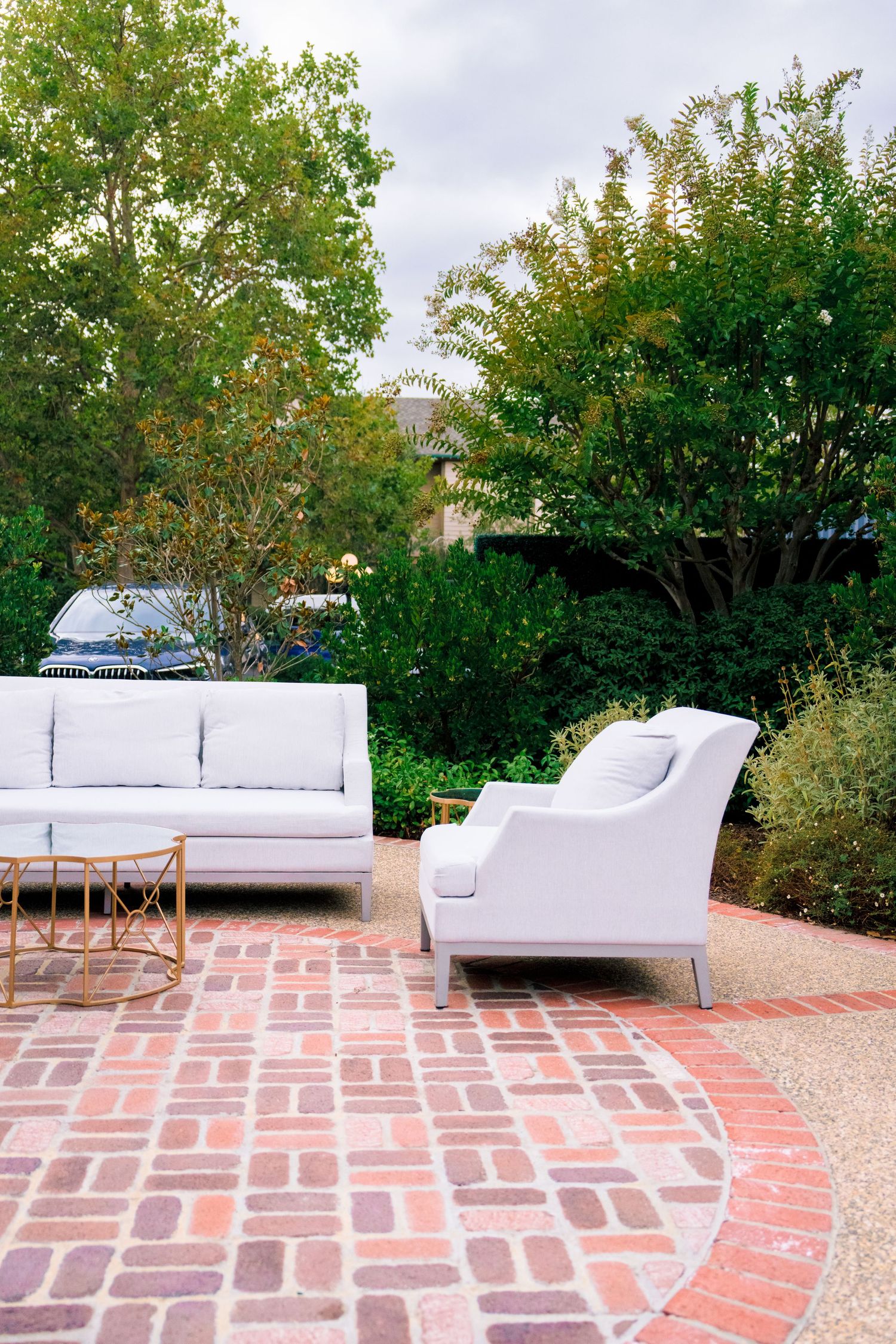 Modern outdoor seating area with white couches on a brick patio surrounded by lush greenery.