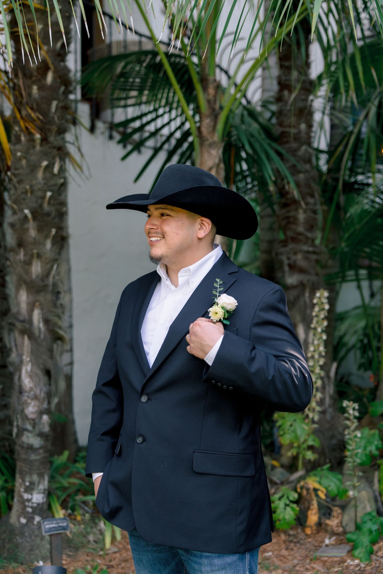 Person in formal black suit and cowboy hat smiling in a garden setting.