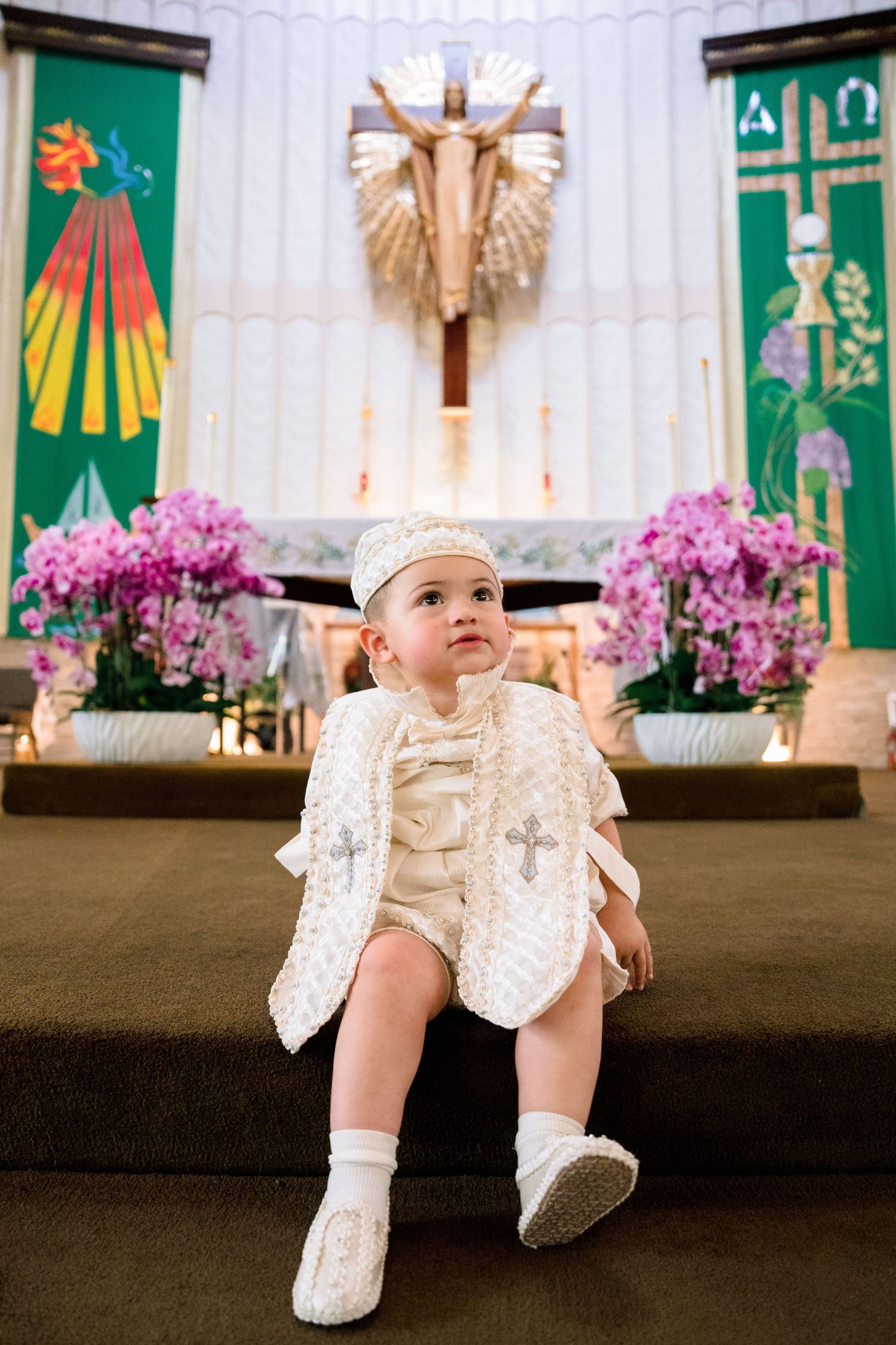 A baby in white christening outfit sits on church altar steps with cross and pink flowers behind.