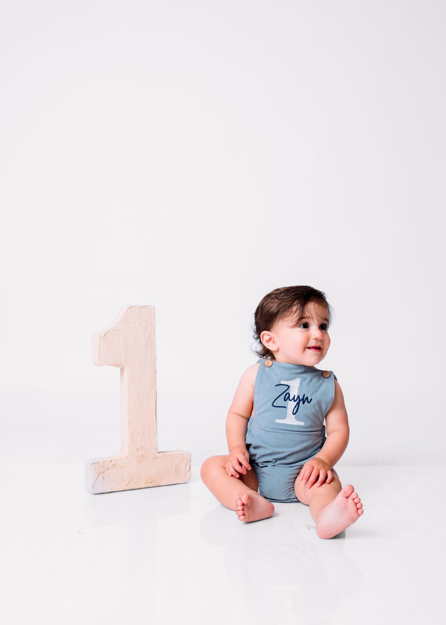 Baby named Zayn sits beside a wooden number 1, celebrating his first birthday in a blue romper.