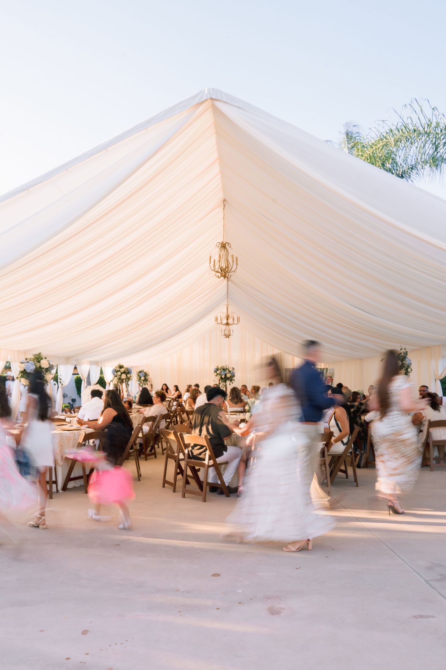 Elegant outdoor wedding reception under a white draped tent with guests mingling and warm chandelier lighting at dusk.