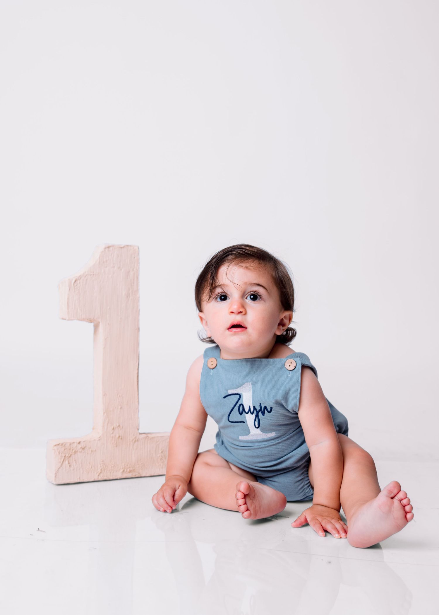 Baby boy named Zayn sits beside a wooden number one, wearing a blue romper, celebrating his first birthday.