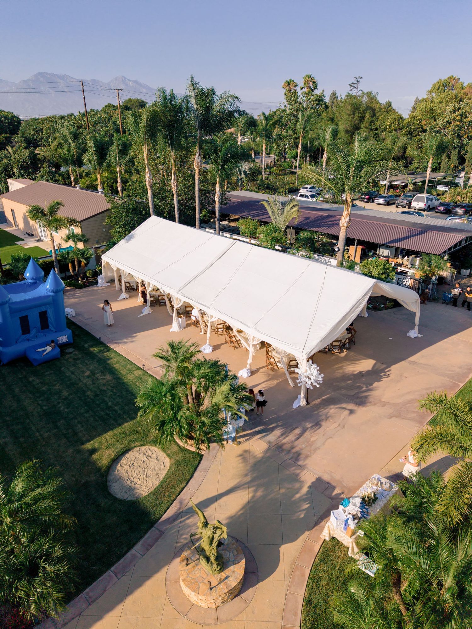 Aerial view of a large white event tent surrounded by palm trees and lush greenery at an outdoor venue.