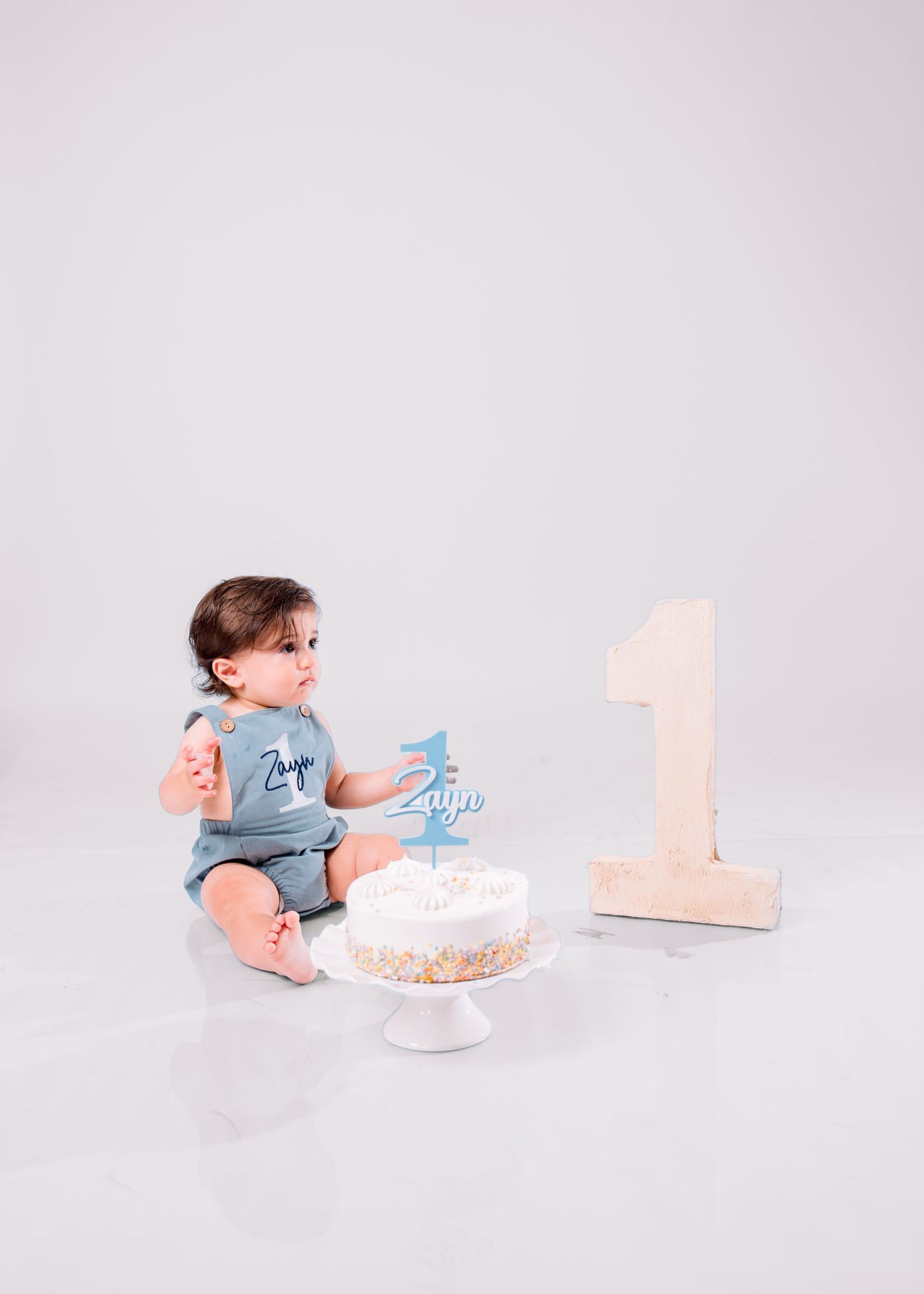 Baby boy named Zayn sits beside a white smash cake and large number 1 decor during his first birthday photo session.