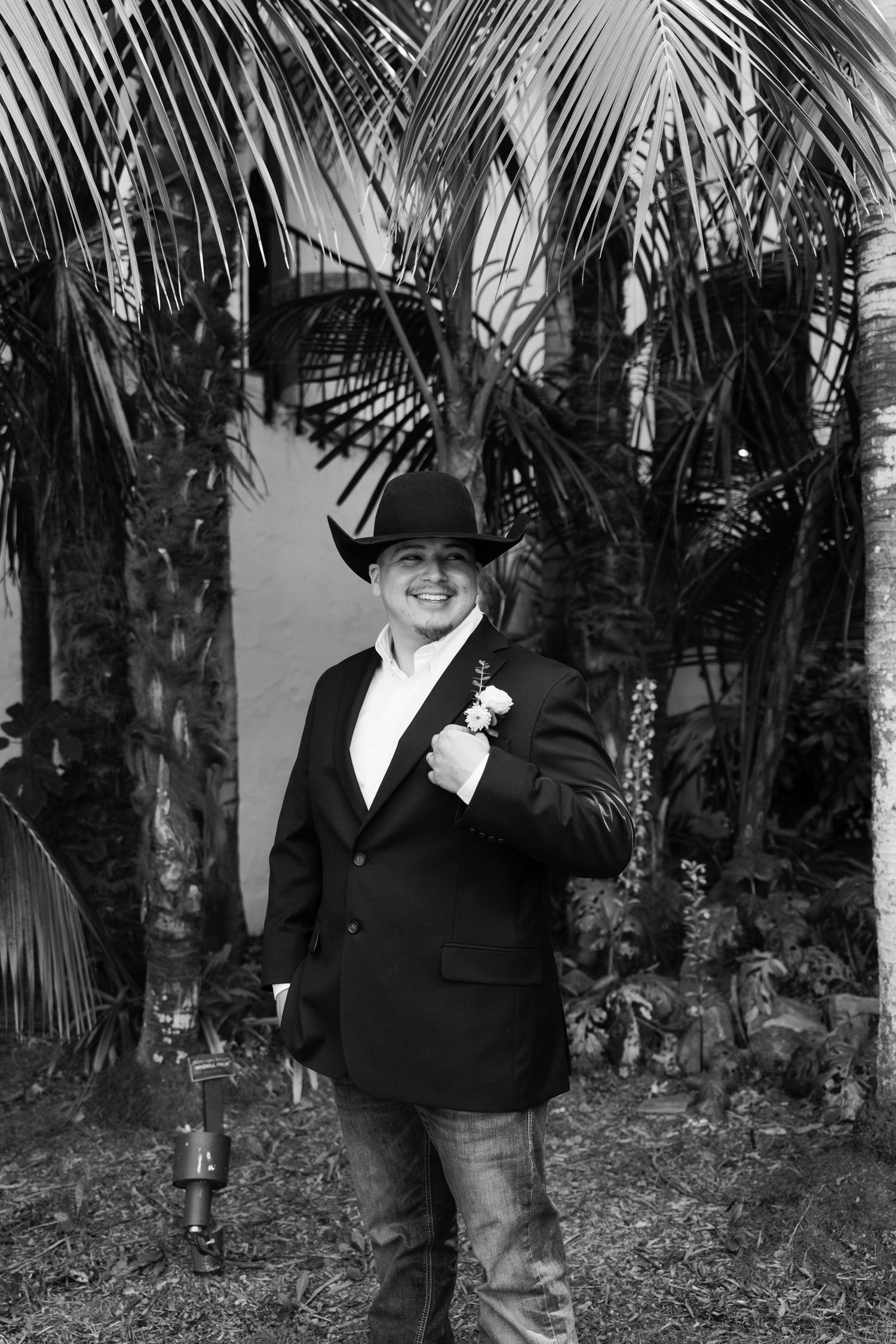Wedding party member in black suit and cowboy hat poses in front of palm trees.