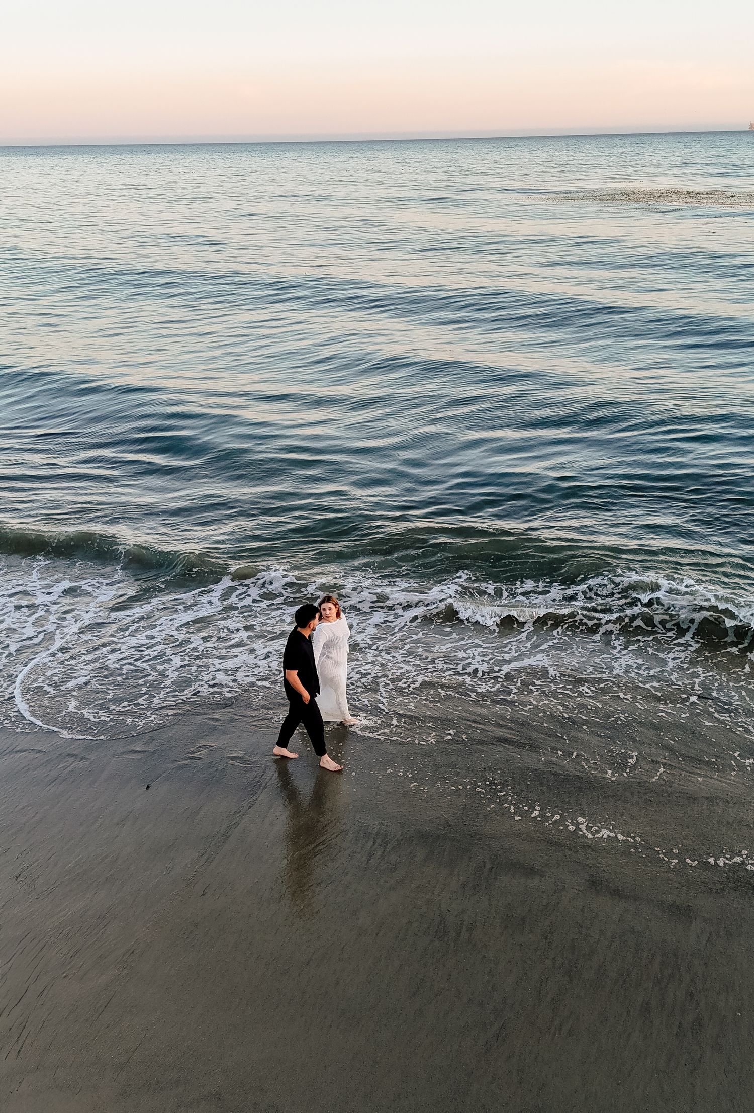 Two people embrace at the shoreline of a calm beach during golden hour sunset.