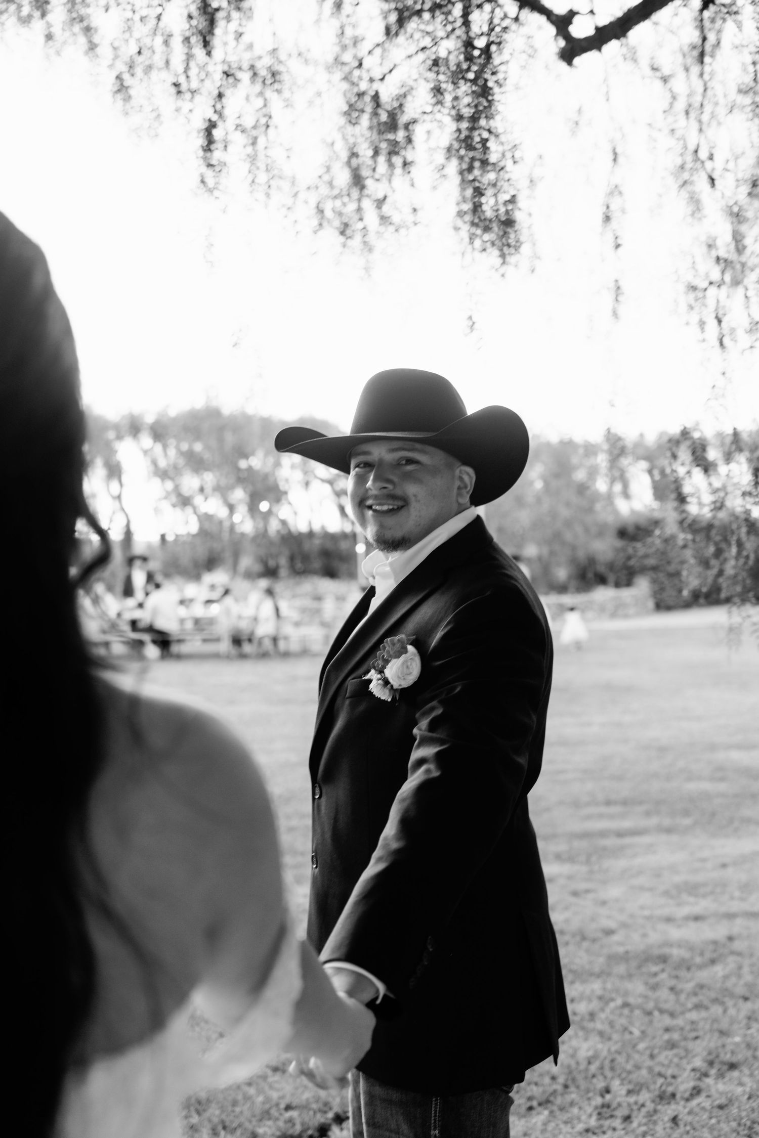 A groom in a black suit and cowboy hat smiles during an outdoor wedding ceremony.