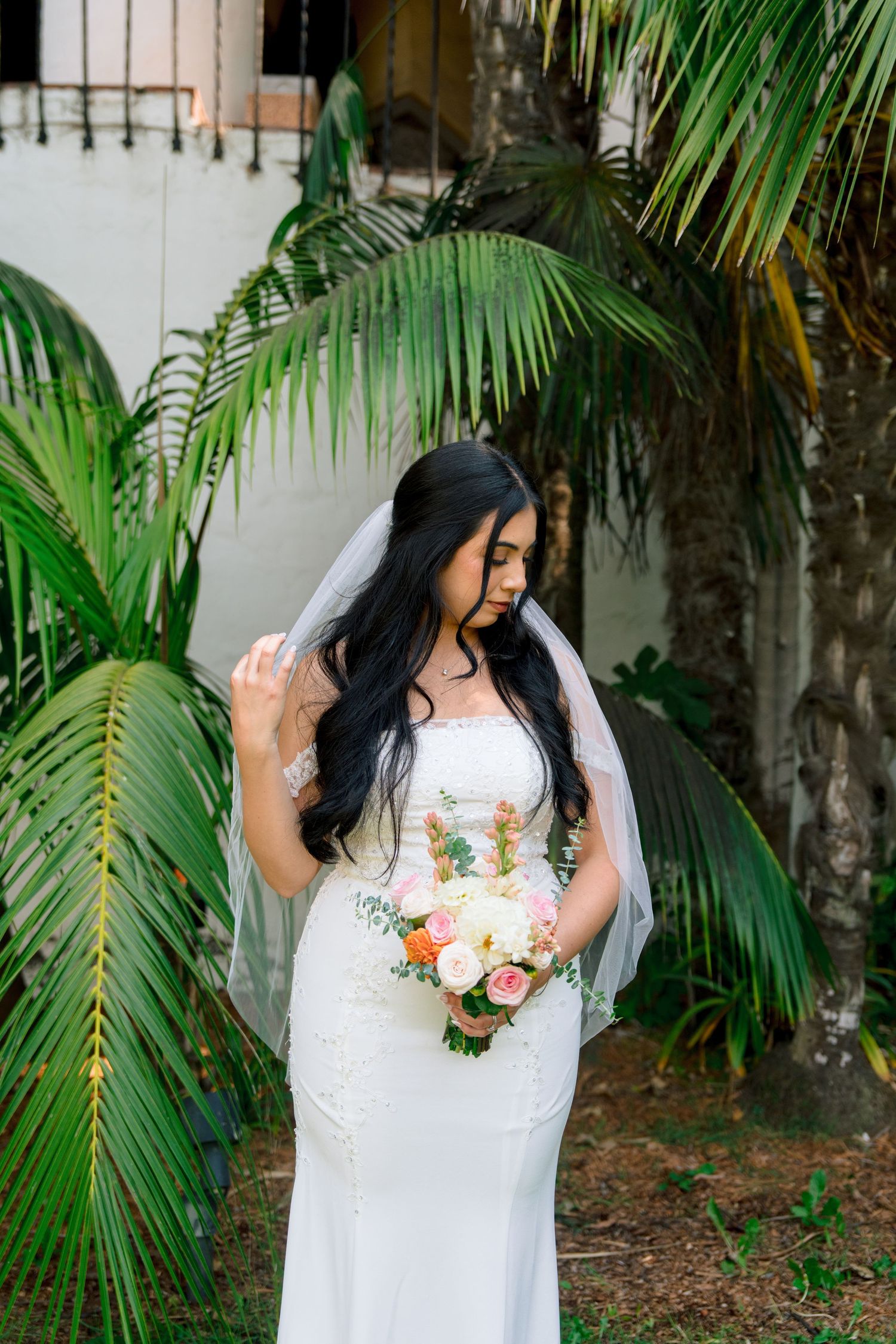 Bride holding bouquet of peach and white flowers poses against palm trees in garden setting.