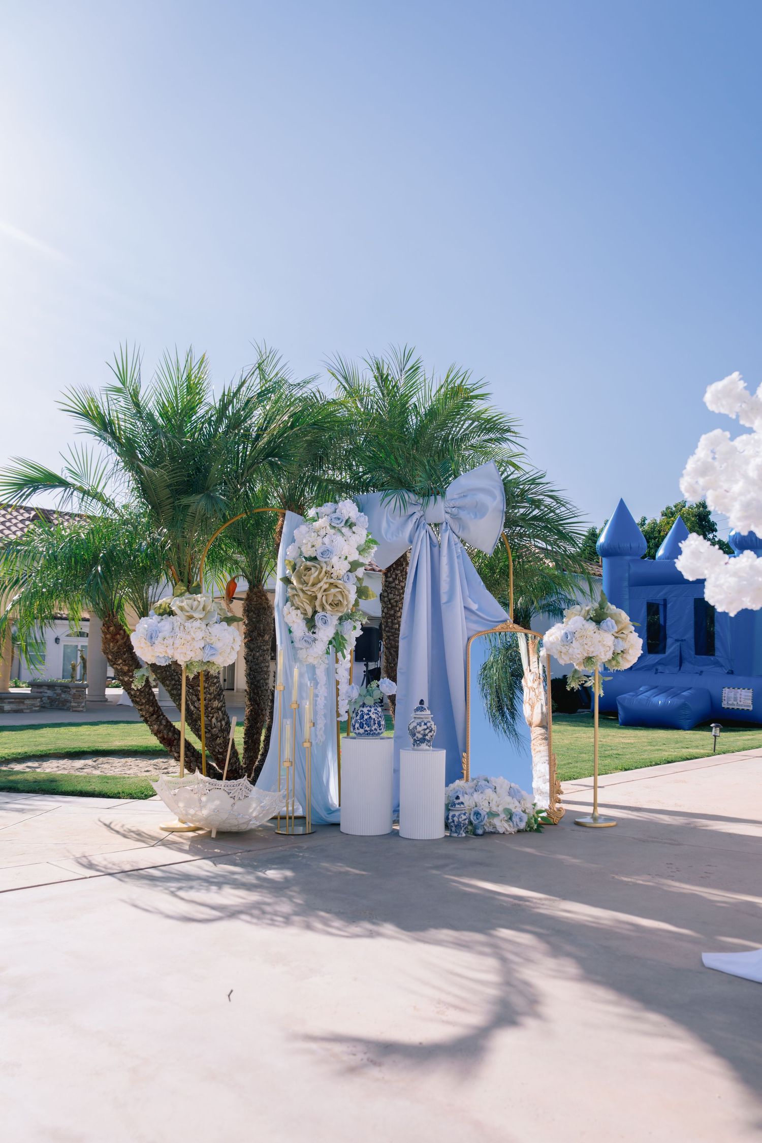 Elegant outdoor wedding arch with blue draping, white flowers, and palm trees under a clear blue sky.