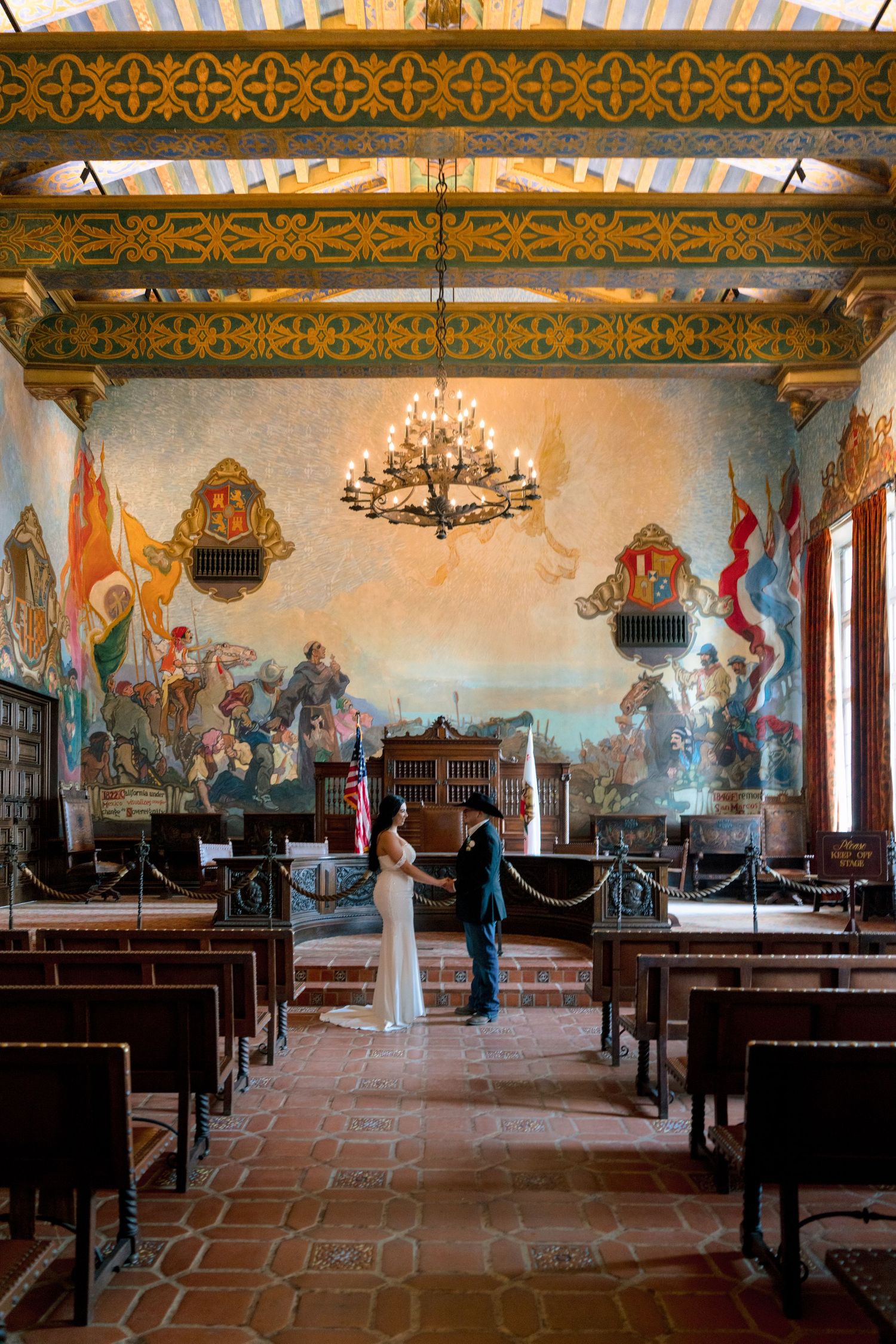 Historic mission room with painted murals, wooden pews and chandeliers.