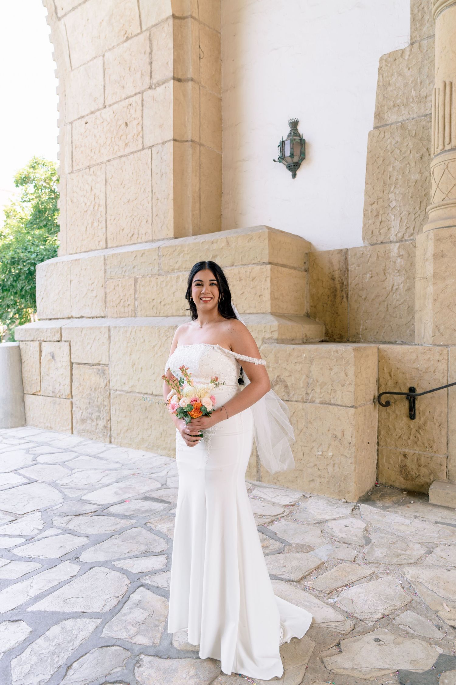 Bride in white off-shoulder dress poses in front of stone archways and columns.