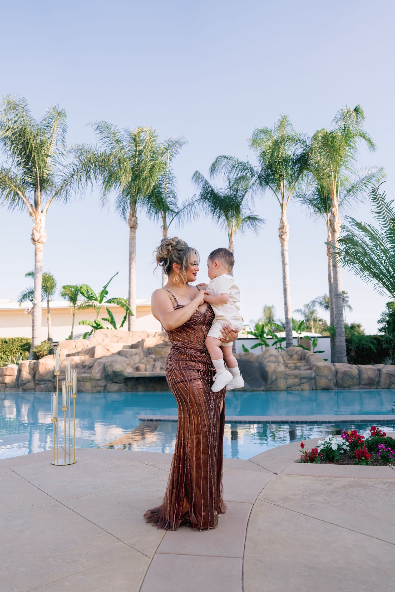 Woman in sequin gown holds baby by poolside with palm trees at sunset.