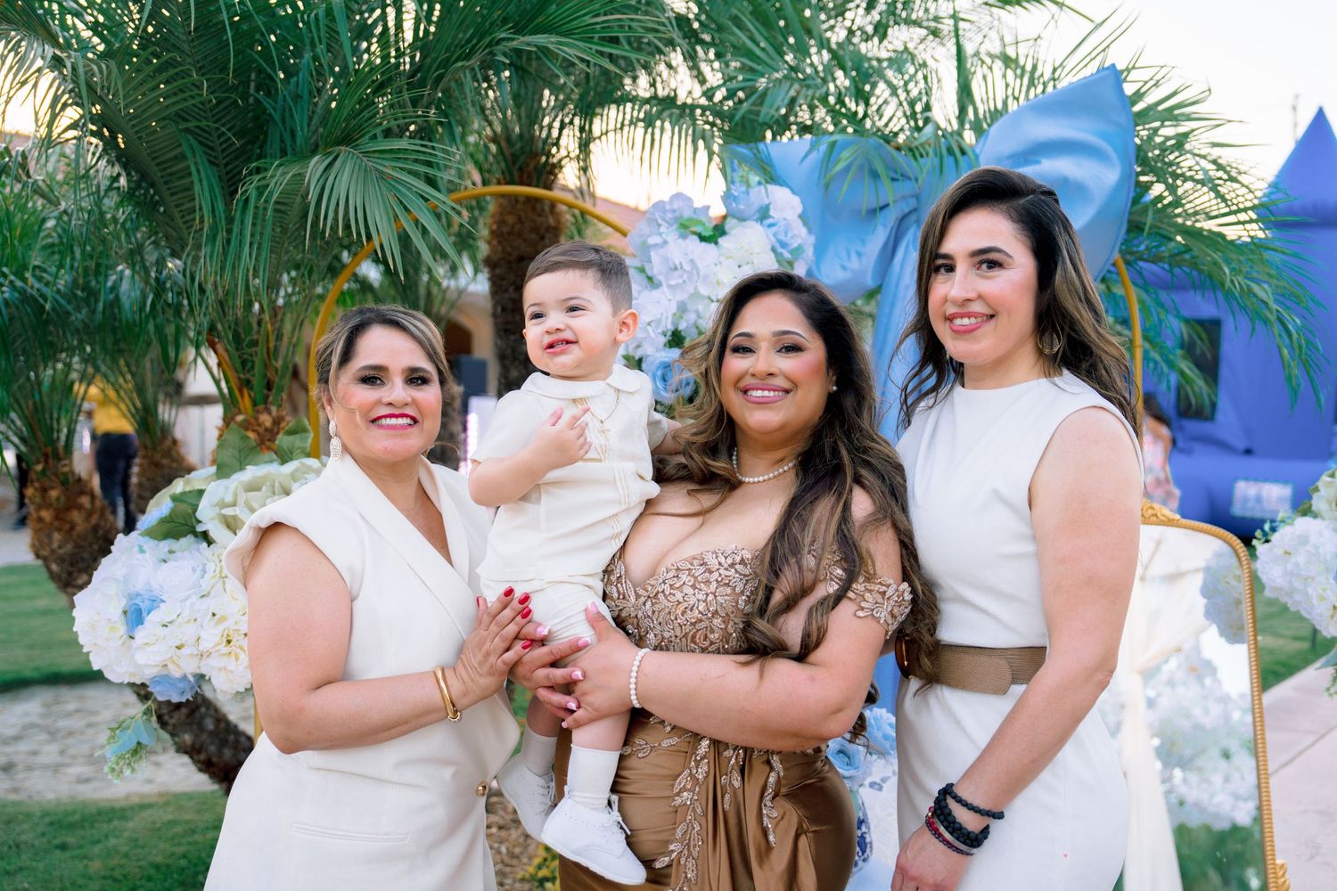 Four women pose outdoors at a blue-themed celebration, one holding a baby dressed in white.