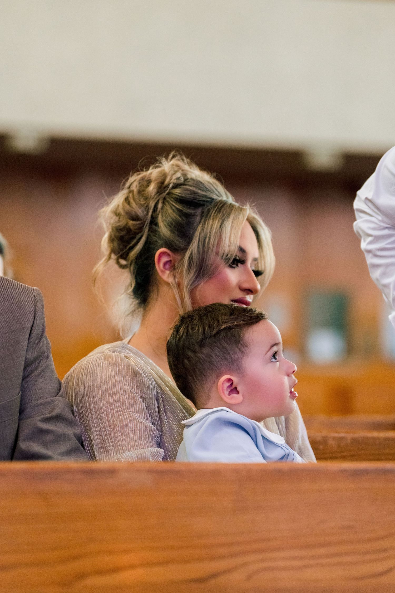 Woman with updo hairstyle sitting in church pew with young boy in light blue shirt during a ceremony.