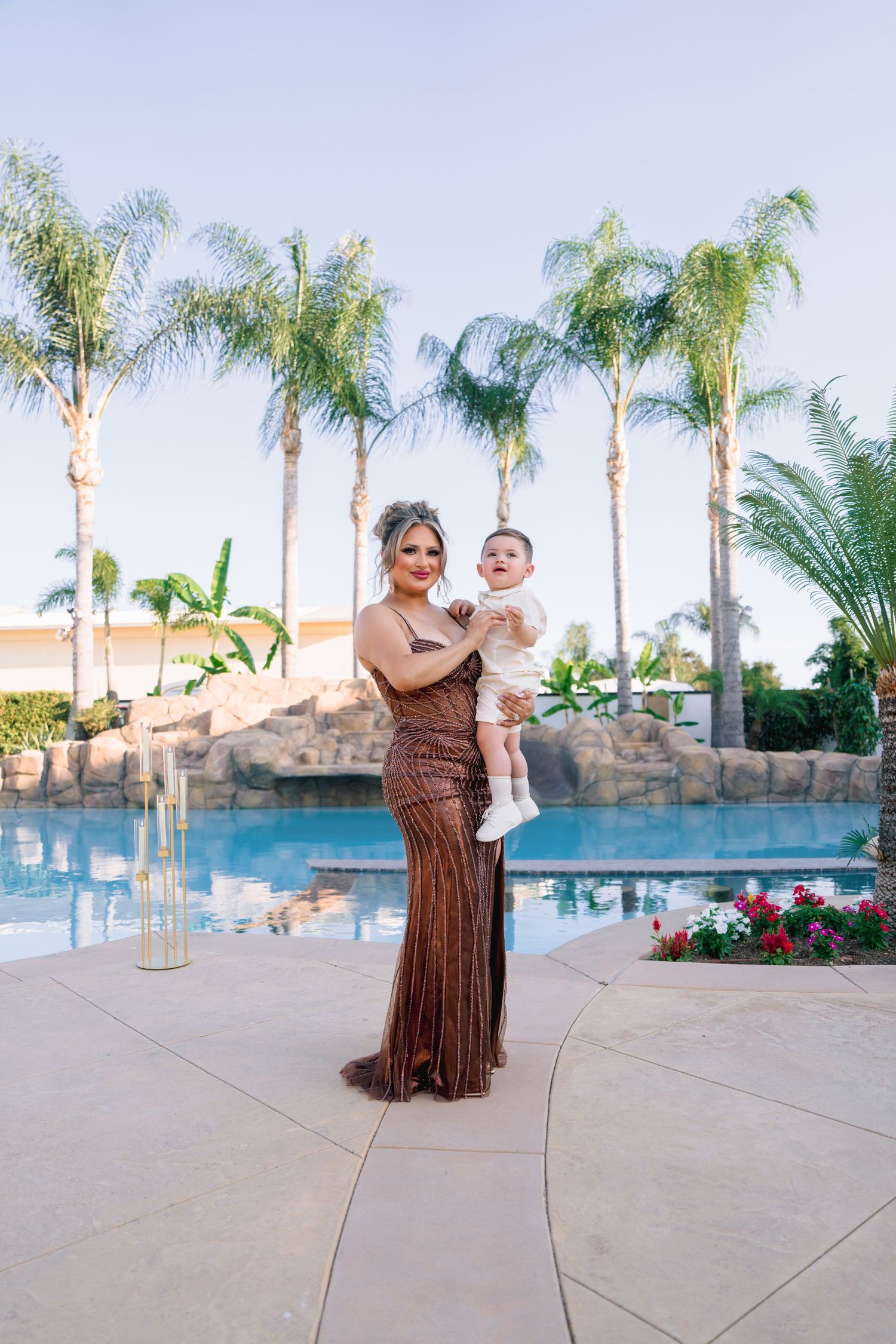 Woman in bronze gown holding baby by resort pool with palm trees.