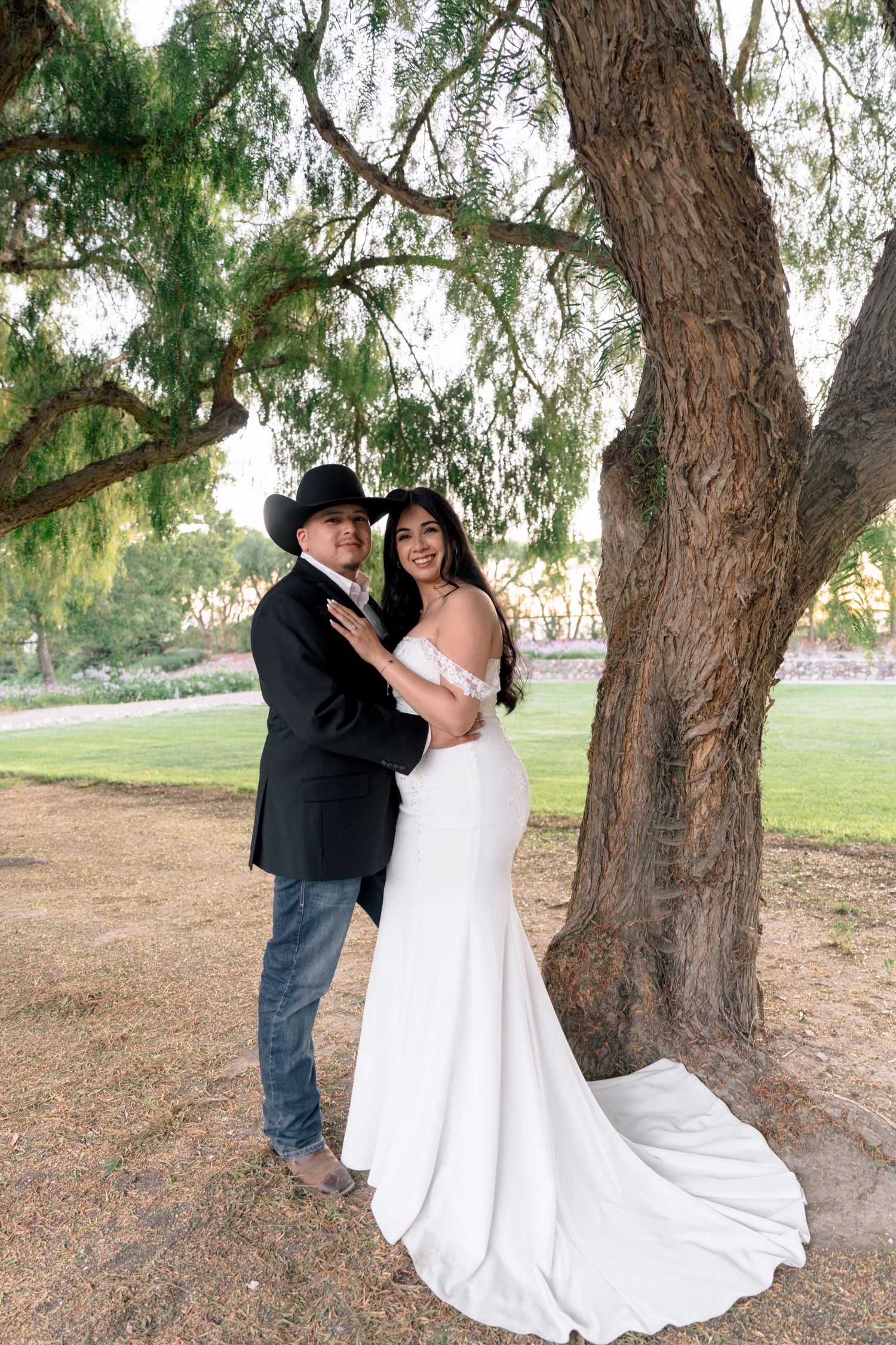 A bride in white gown and groom in cowboy attire pose together under a large tree.