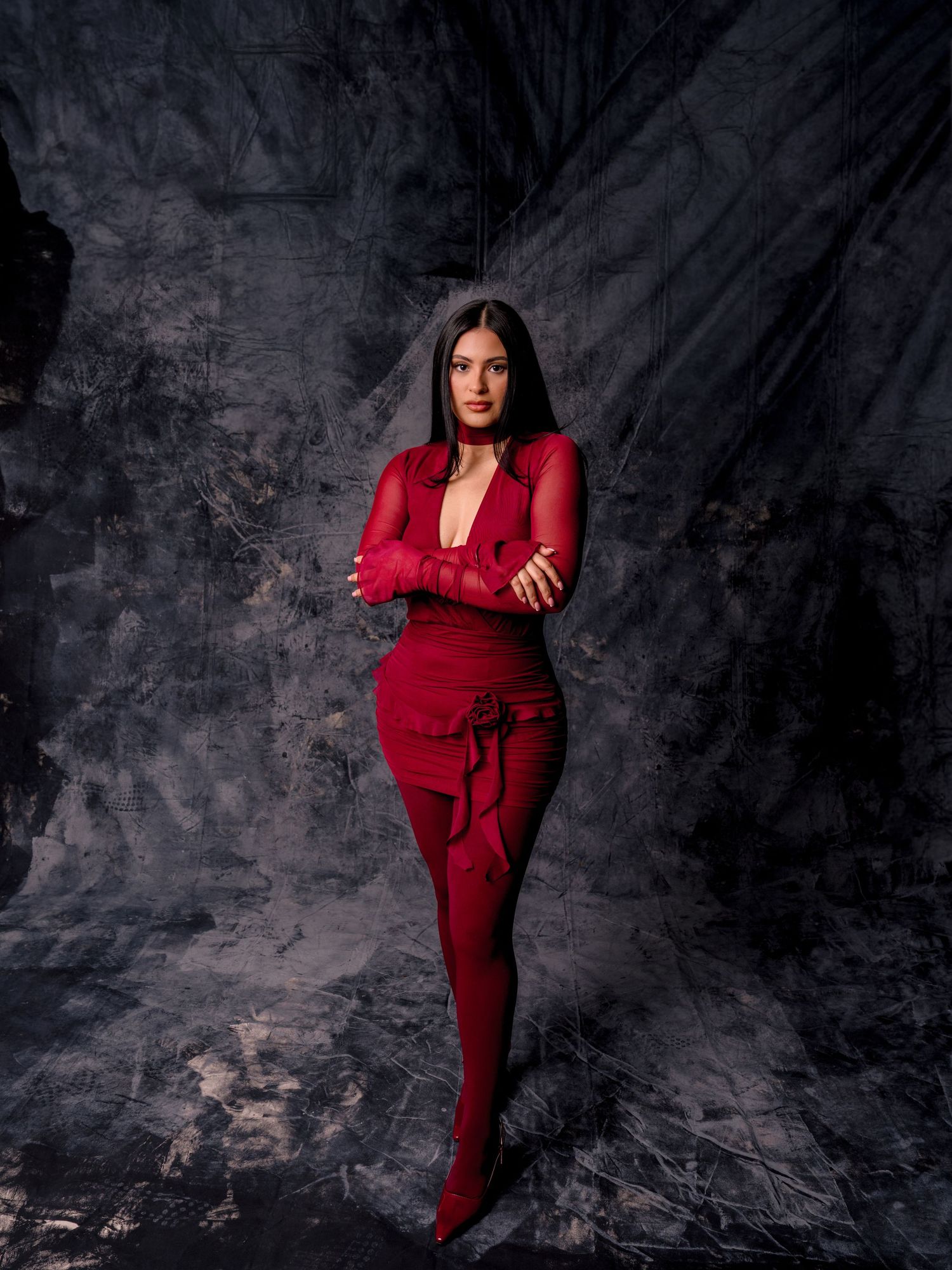 Woman in red dress, tights, and heels posing confidently against dark textured backdrop.