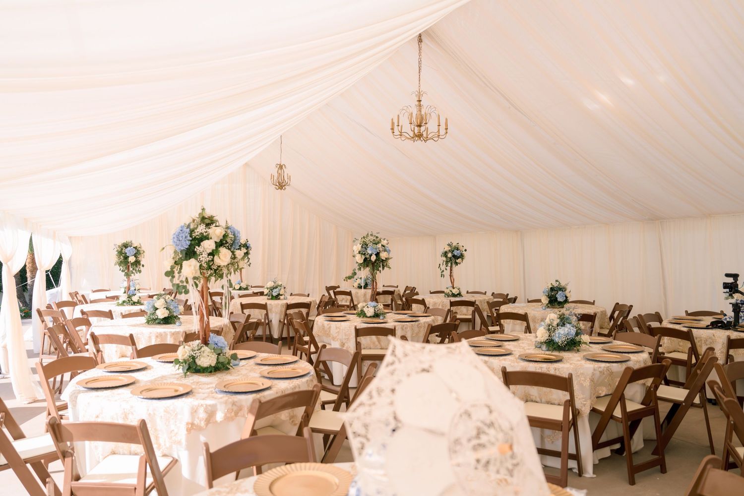 Elegant wedding reception tent with floral centerpieces, draped white fabric ceiling, round tables, and wooden folding chairs.