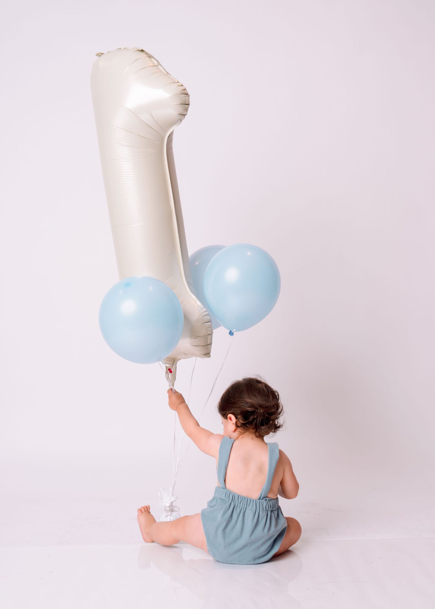 Baby boy sitting on white floor holding light blue balloons and a number one foil balloon on his first birthday.