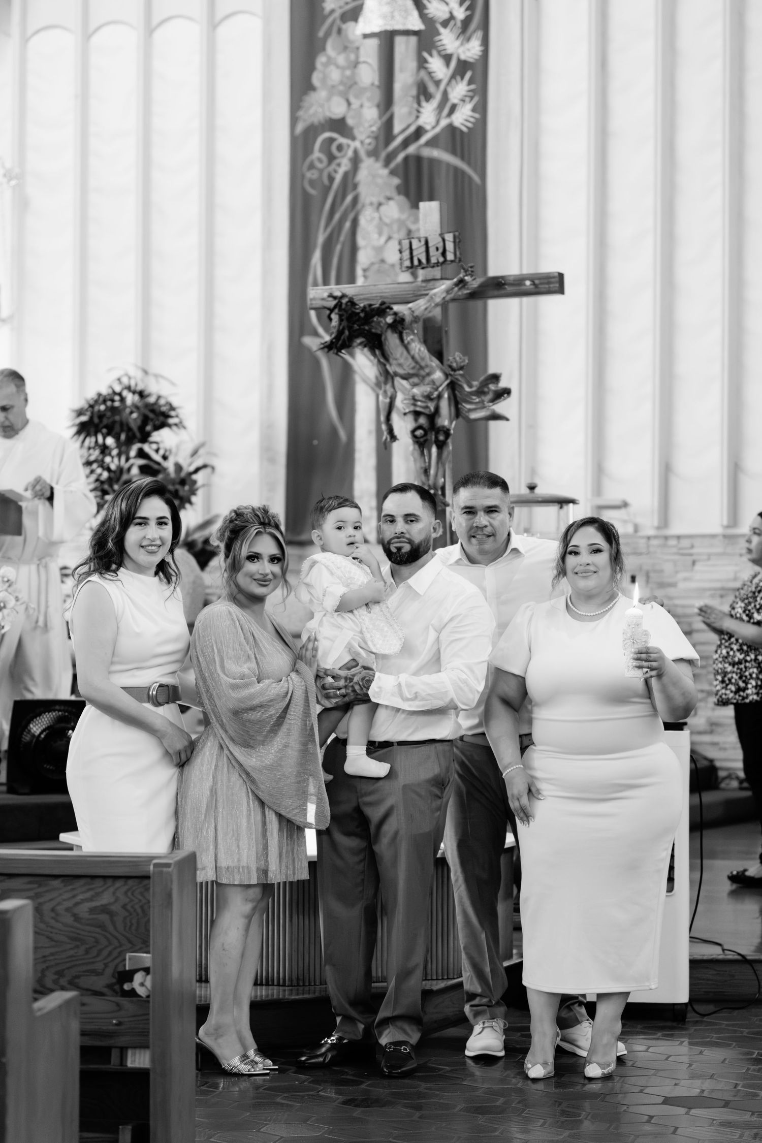 Black and white photo of a family posing together in a church during a baptism ceremony with a crucifix in the background.