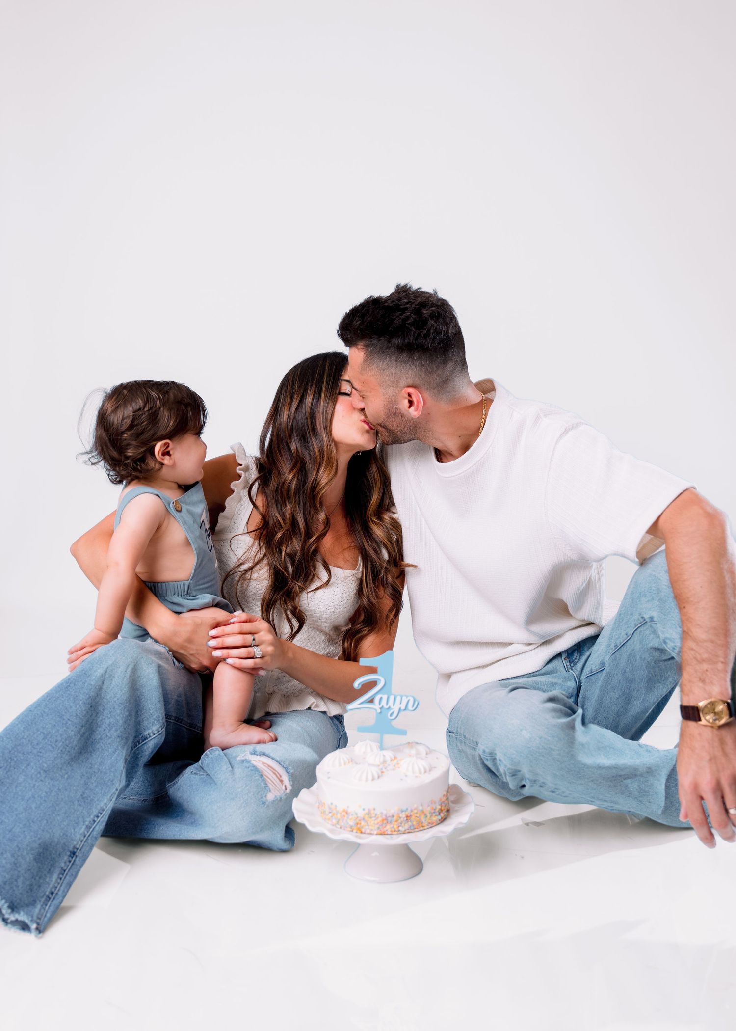 Family celebrating toddler's 2nd birthday with a small cake, parents kissing while child sits beside them on white background.