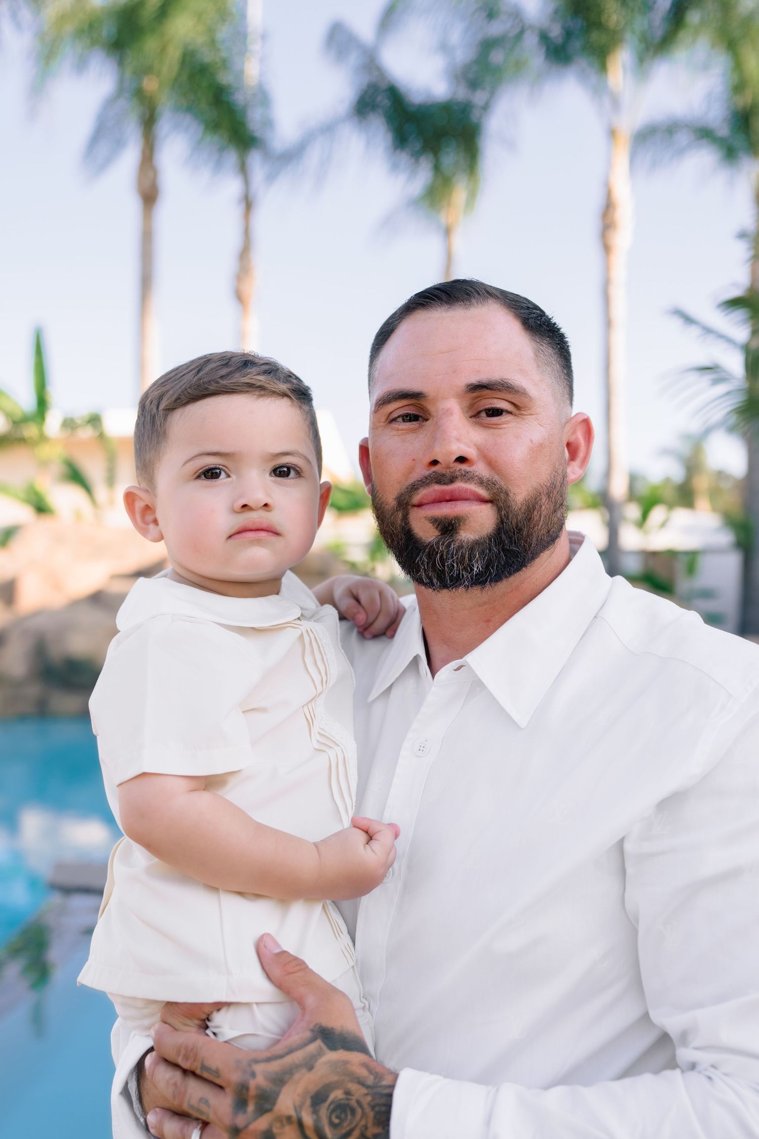 A bearded father with tattoos holds his young son near a pool with palm trees, both wearing white outfits.