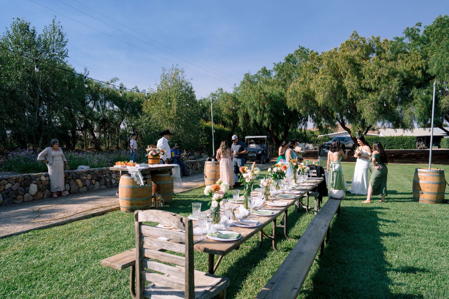 Long outdoor dining table setup with rustic benches on a grassy lawn under string lights at sunset.