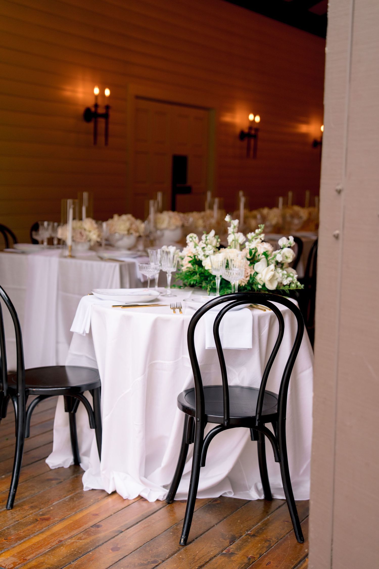 Wedding reception venue with white tablecloths and black bentwood chairs in a rustic indoor setting.